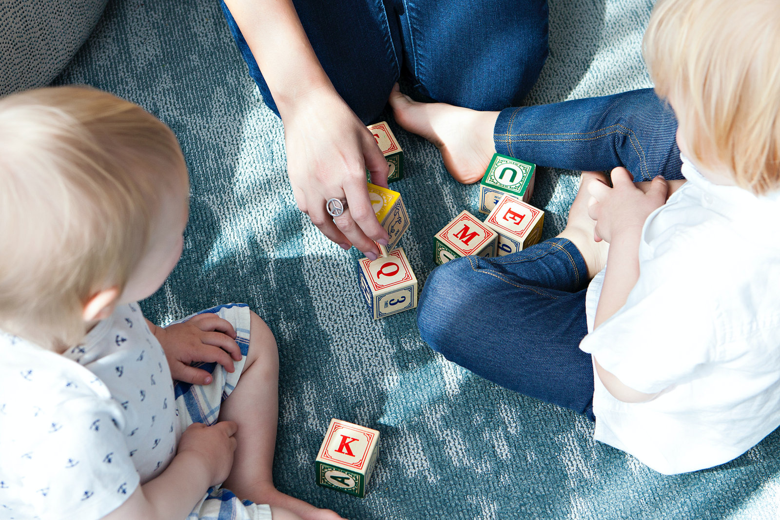 Two toddlers playing with alphabet blocks on a rug beside a caregiver