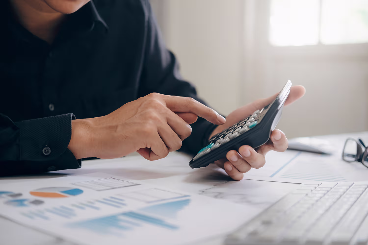 Person wearing a dark shirt using a calculator at a desk with financial charts and a keyboard.