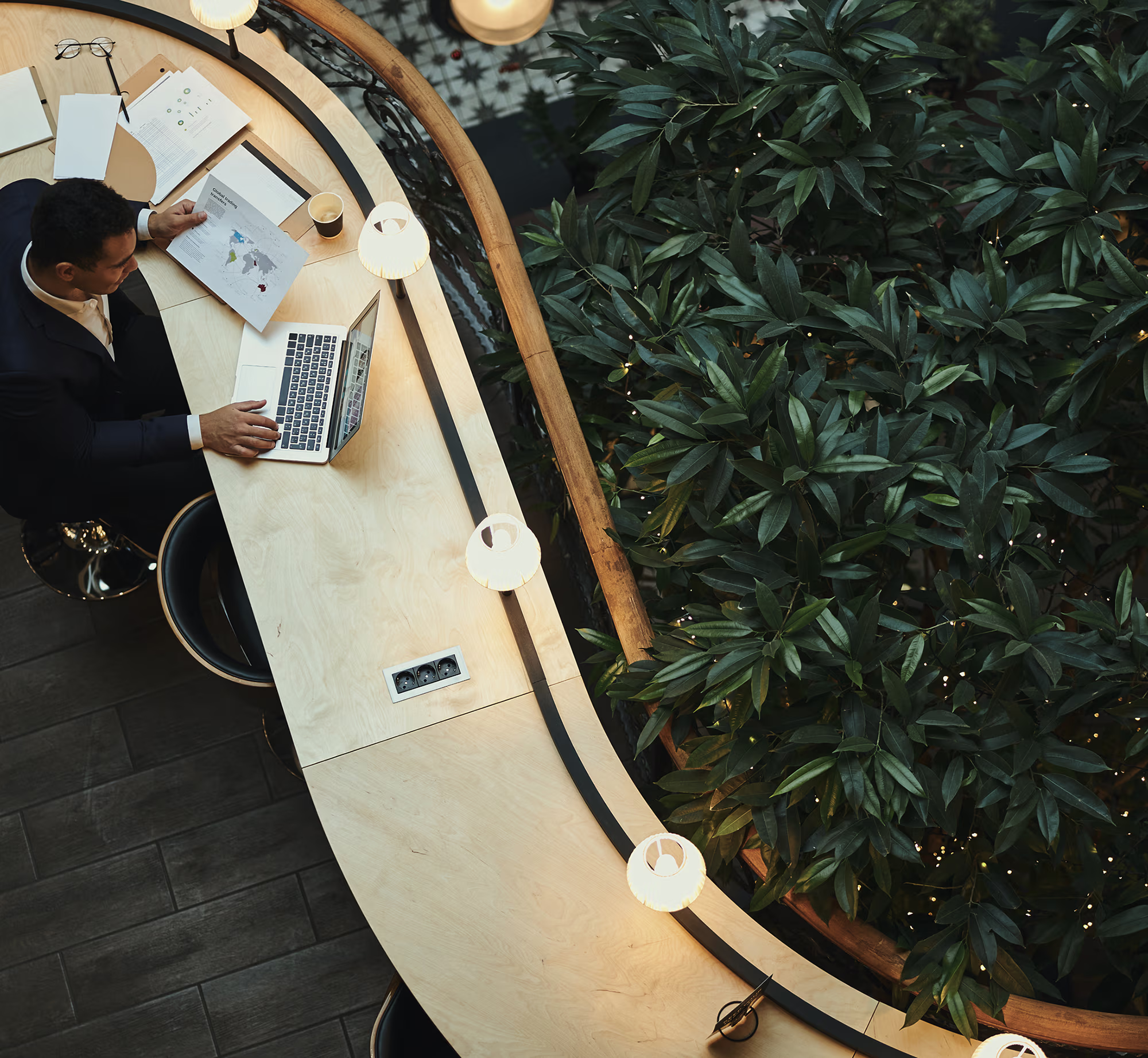 Overhead view of a man in a dark suit working at a curved wooden desk with a laptop, holding a paper with a world map, next to a large green leafy plant and illuminated by small round lamps.