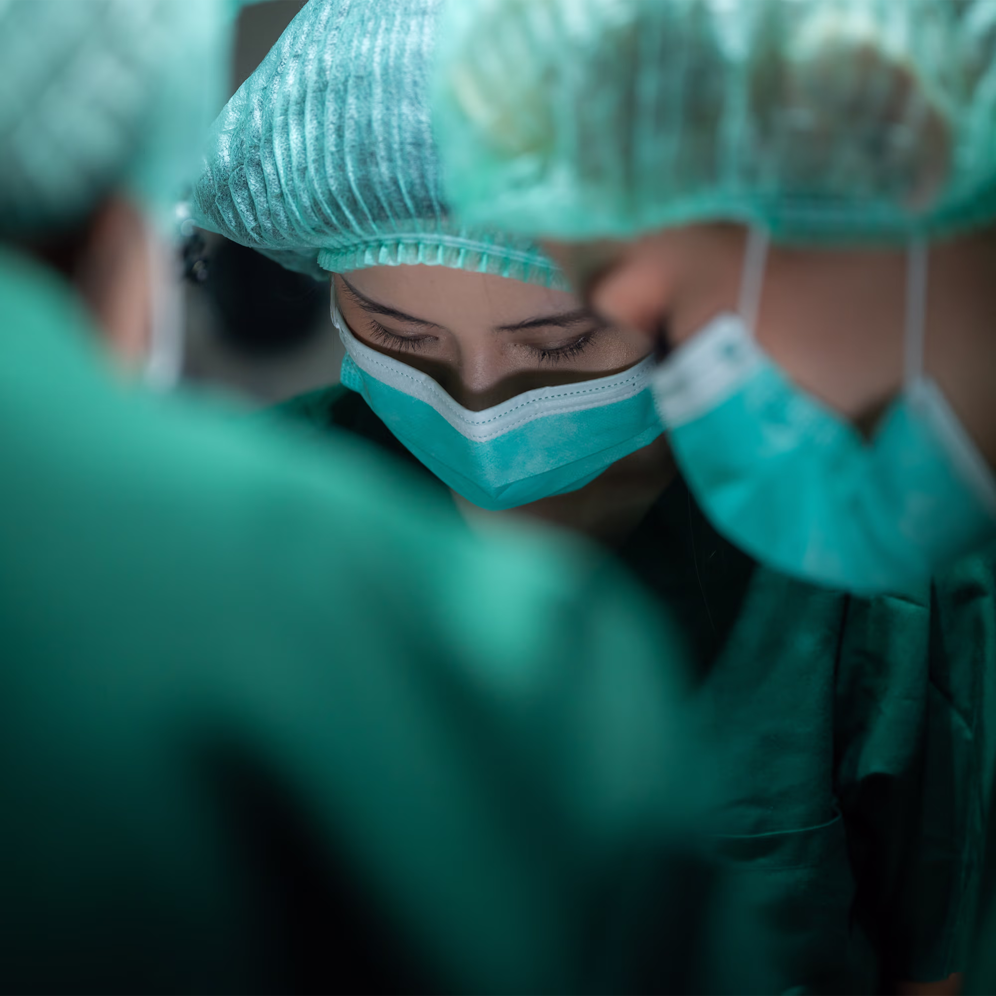Close-up of two healthcare workers wearing surgical masks and hair covers, focused intently, with blurred foreground and background.