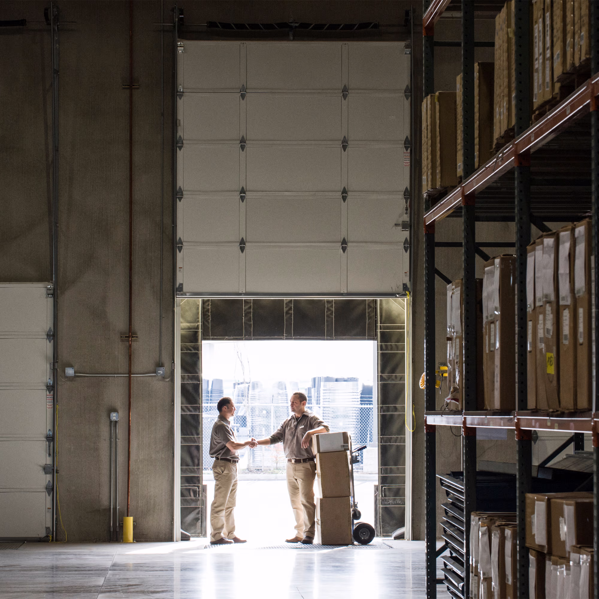 Two men shaking hands inside a warehouse near a large open garage door, with boxes stacked on a hand truck and shelves filled with more boxes to the right.