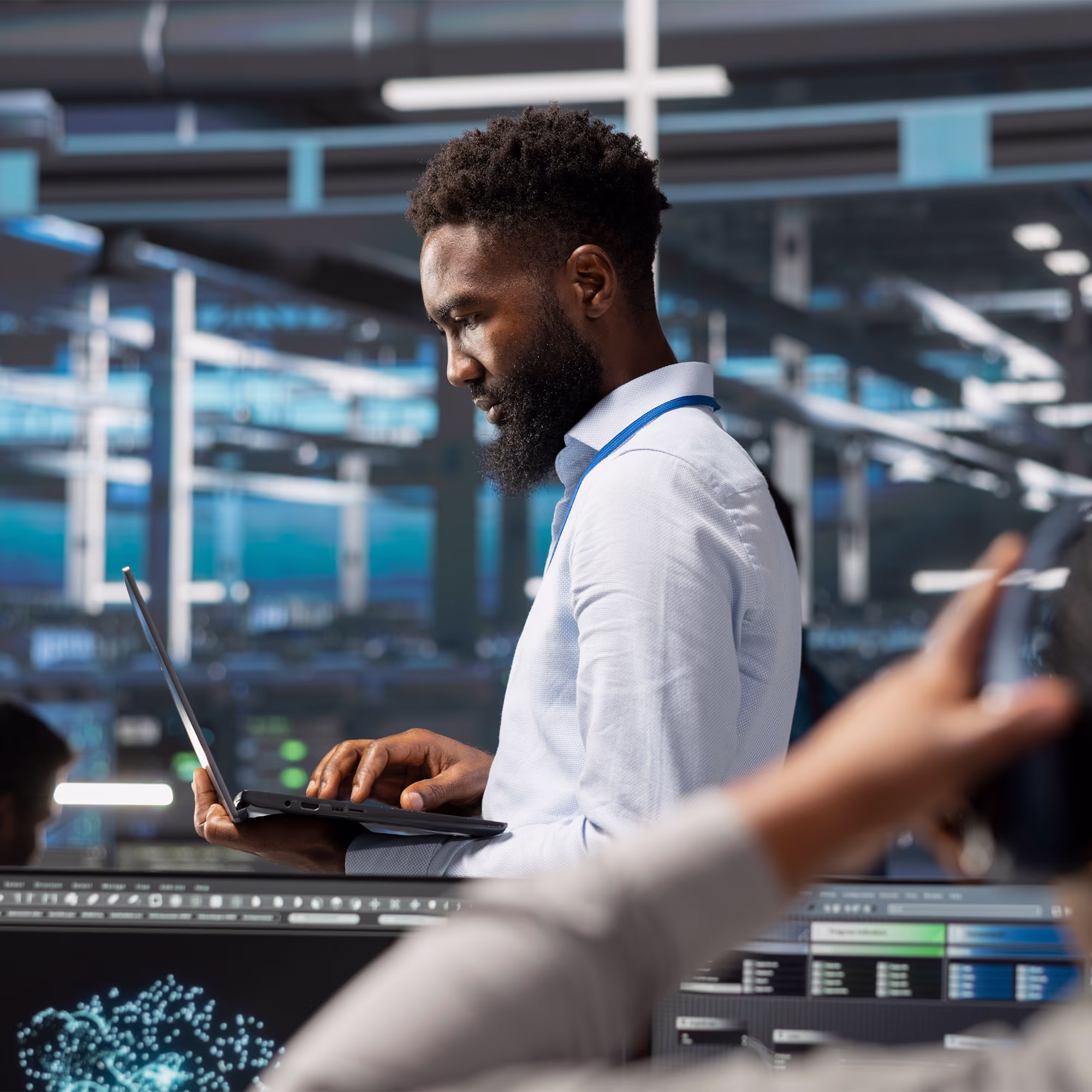 Man with a beard standing indoors using a laptop, behind him is a high-tech workspace with illuminated screens and infrastructure.