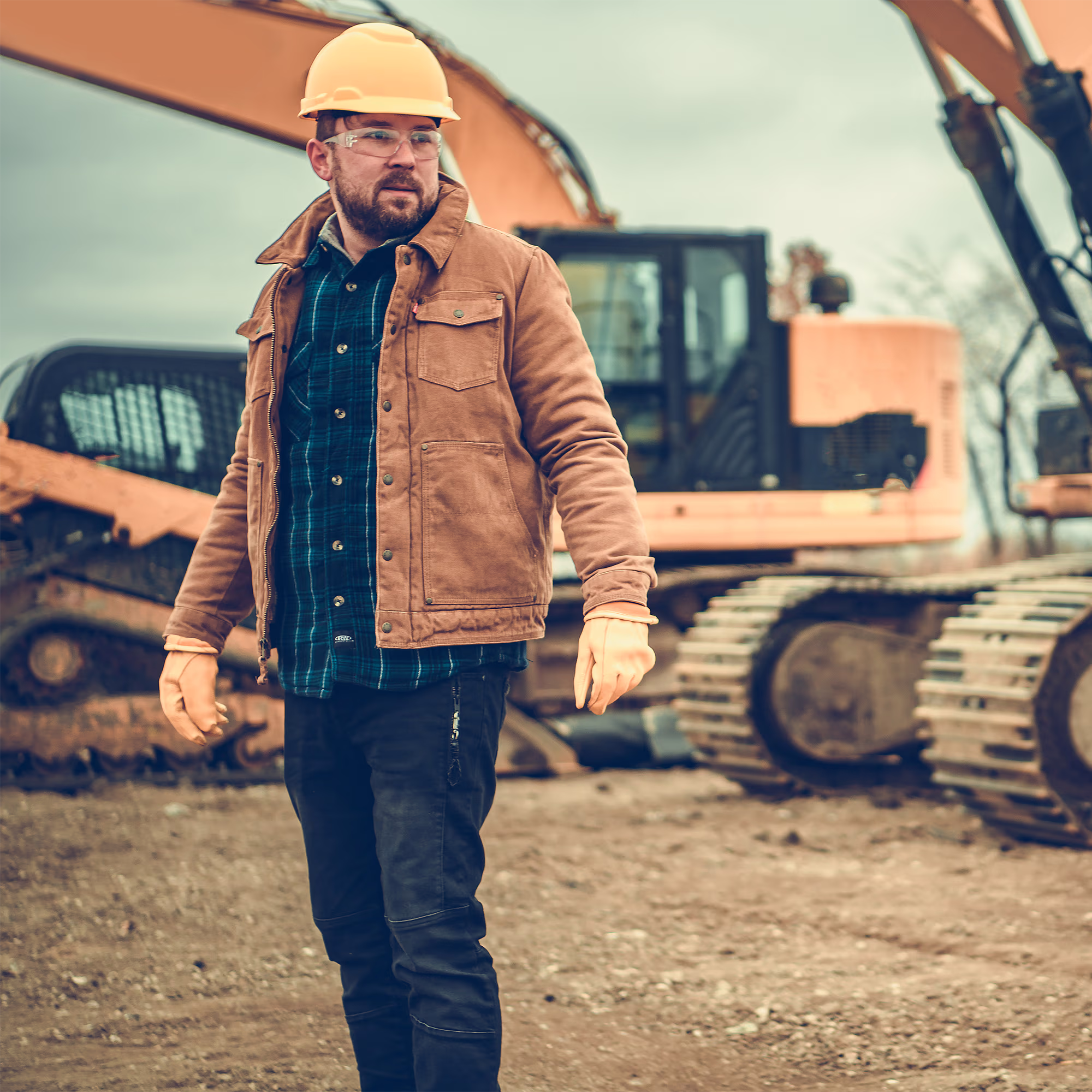Construction worker wearing a yellow hard hat, safety glasses, brown jacket, and gloves standing on a dirt site with orange heavy machinery in the background.