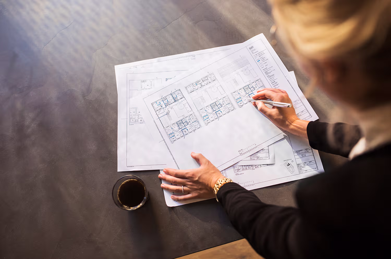 Person in business attire reviewing architectural floor plans on a desk with a cup of coffee nearby.