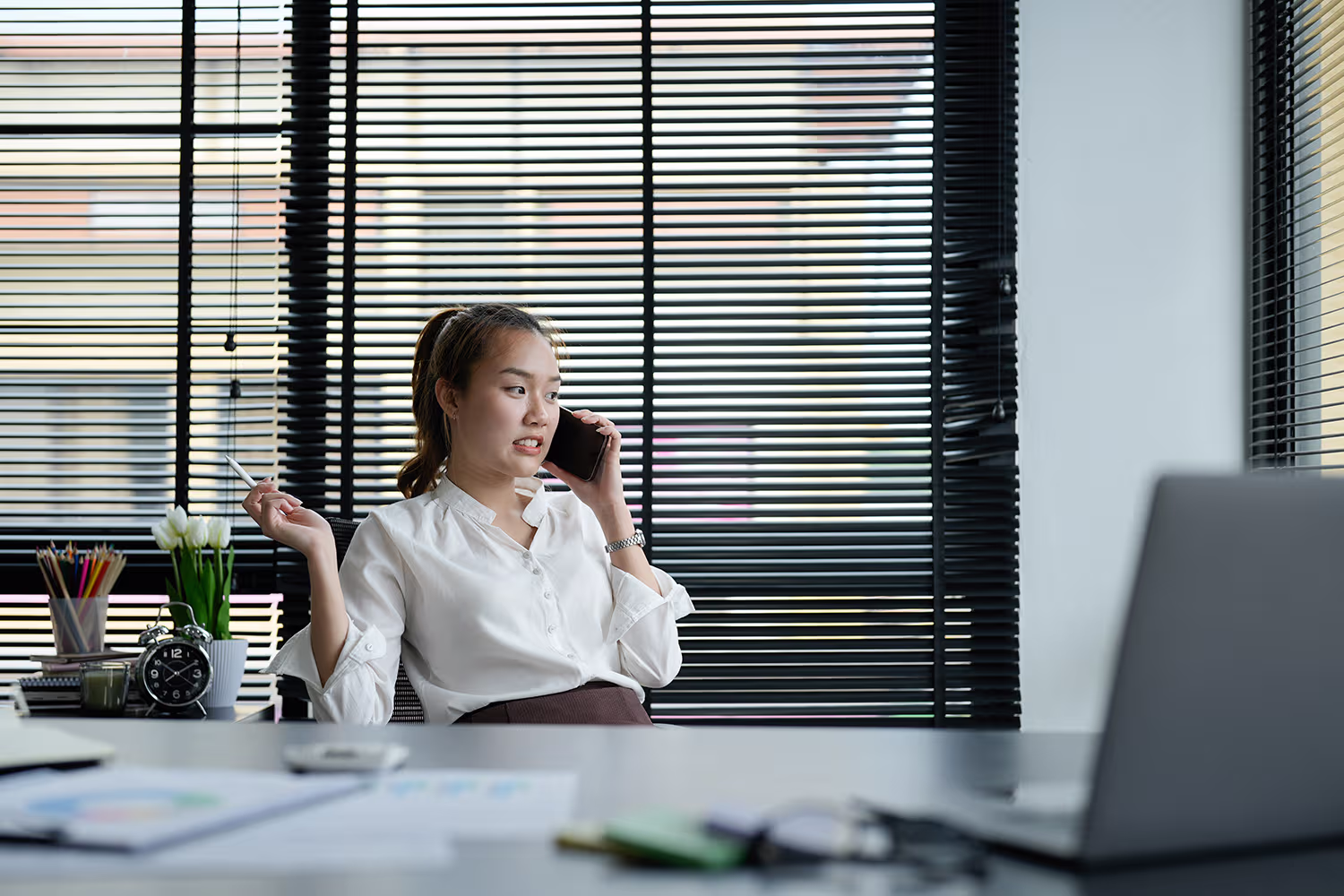 A woman in a white blouse sitting at a desk in an office with black blinds behind her, talking on a phone and holding a pen, with a laptop and office supplies on the desk.