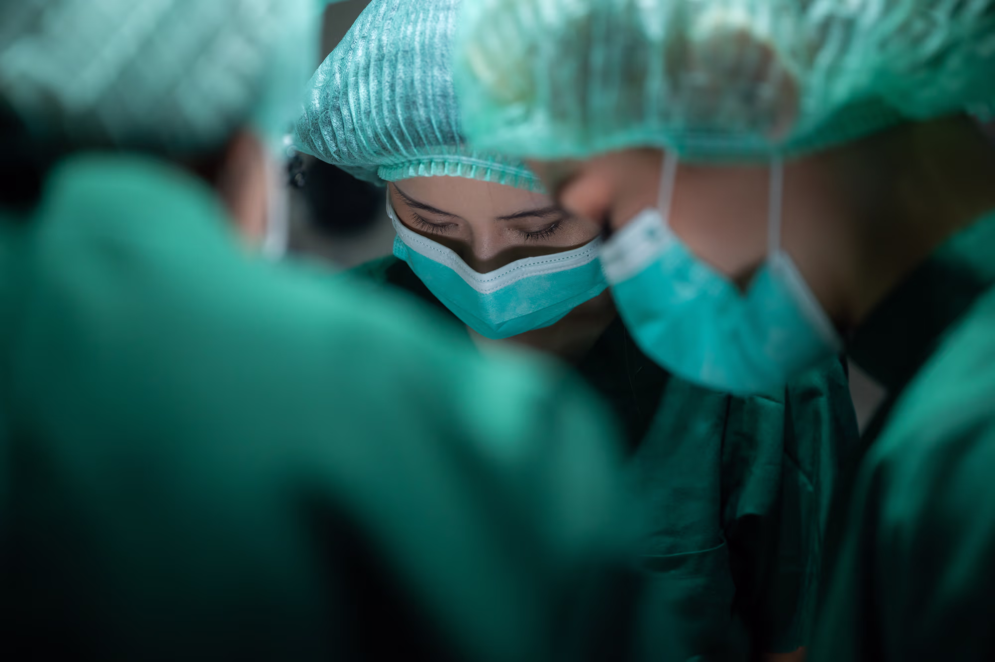 Three medical professionals wearing surgical masks and green scrubs focused on a task, with one person in the center looking downward.