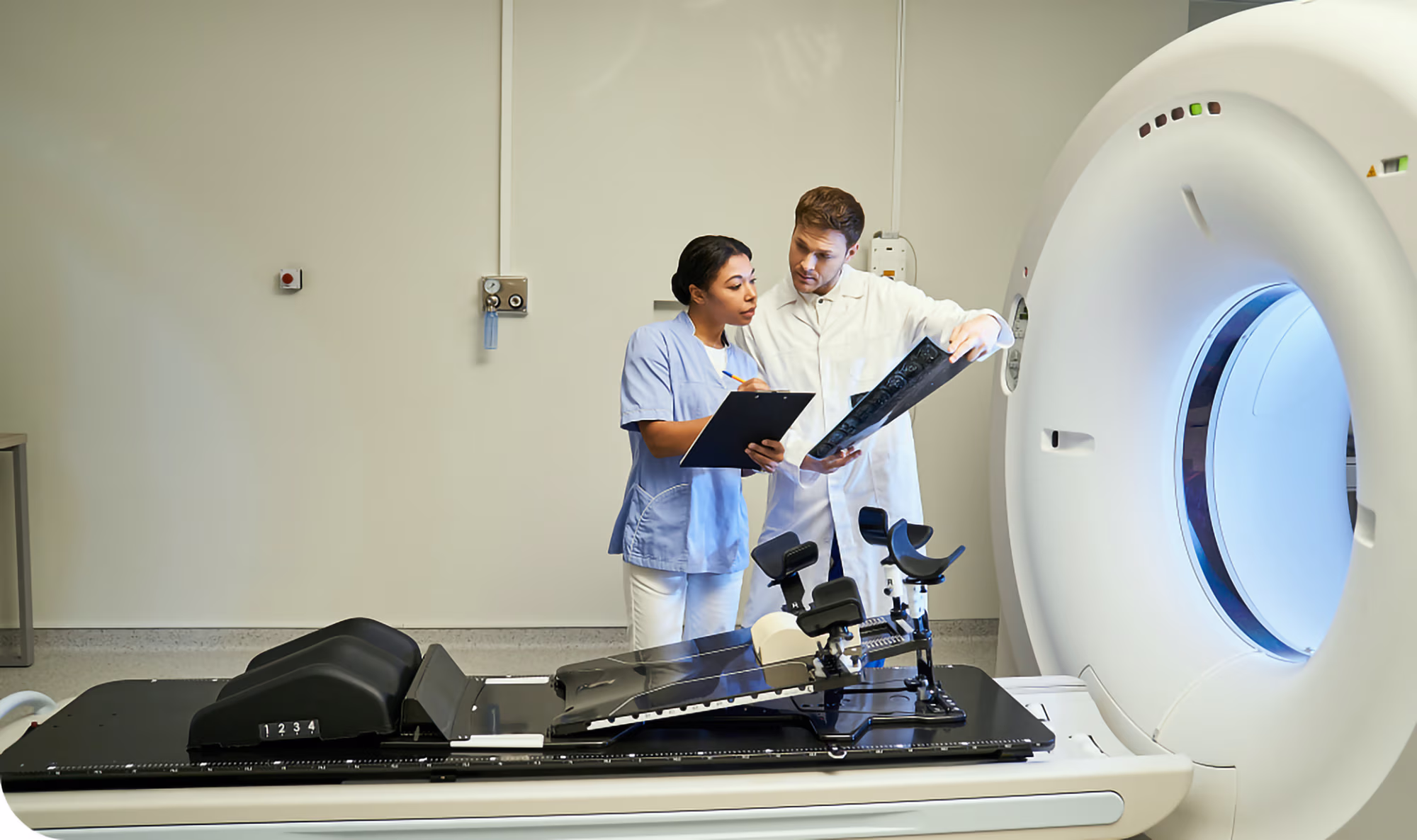 Two medical professionals in a clinical room reviewing a patient scan beside a large CT or MRI scanner machine.