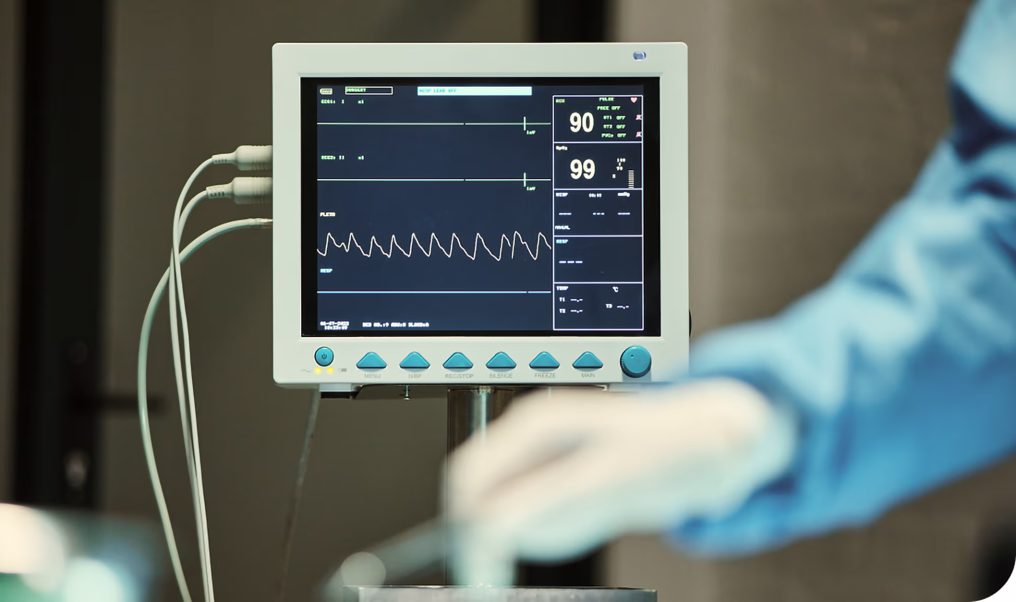 Hospital patient monitor displaying a heart rate of 90 and oxygen saturation at 99 percent with waveforms, attached cables visible on the left and blurred gloved hand in blue scrubs on the right.