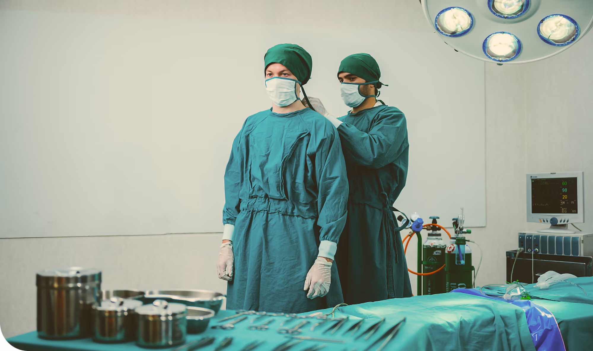 Two medical professionals in green surgical gowns and masks prepare in an operating room, with surgical tools and equipment on a covered table nearby.