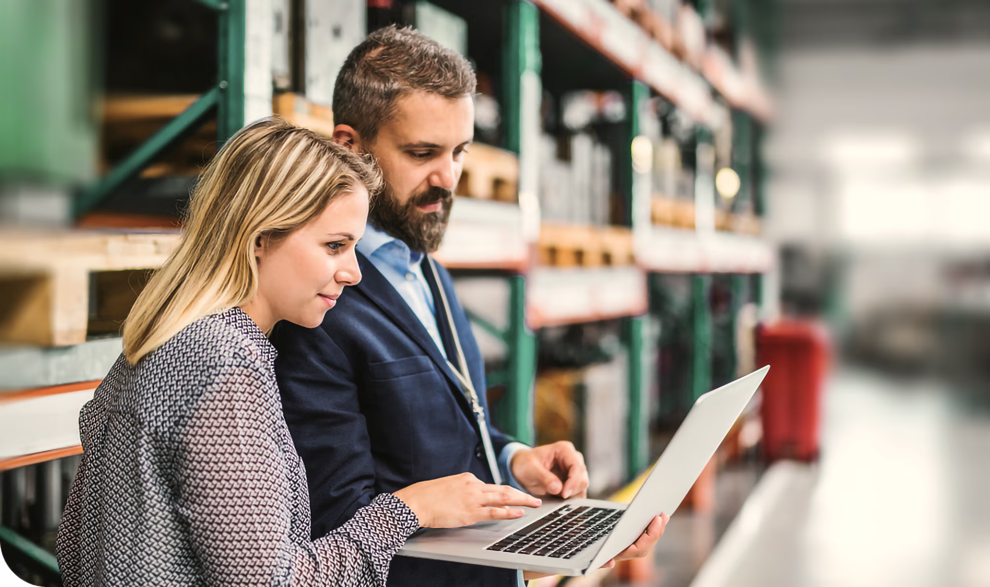 A man and woman standing in a warehouse are looking at a laptop together, with shelves stocked with boxes and pallets in the background.