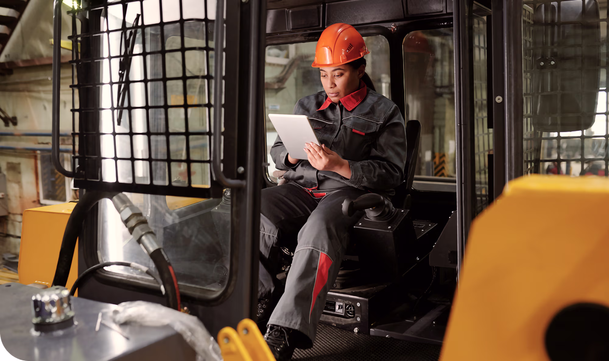 Worker wearing an orange safety helmet and dark work uniform sitting inside industrial machinery cab, reviewing a tablet device.