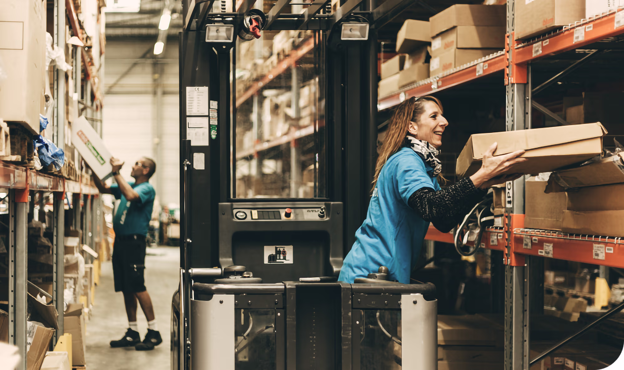 Two warehouse workers in blue shirts organizing and retrieving boxes from shelves in an industrial storage aisle, with one handling a cardboard box and the other reaching for an item.