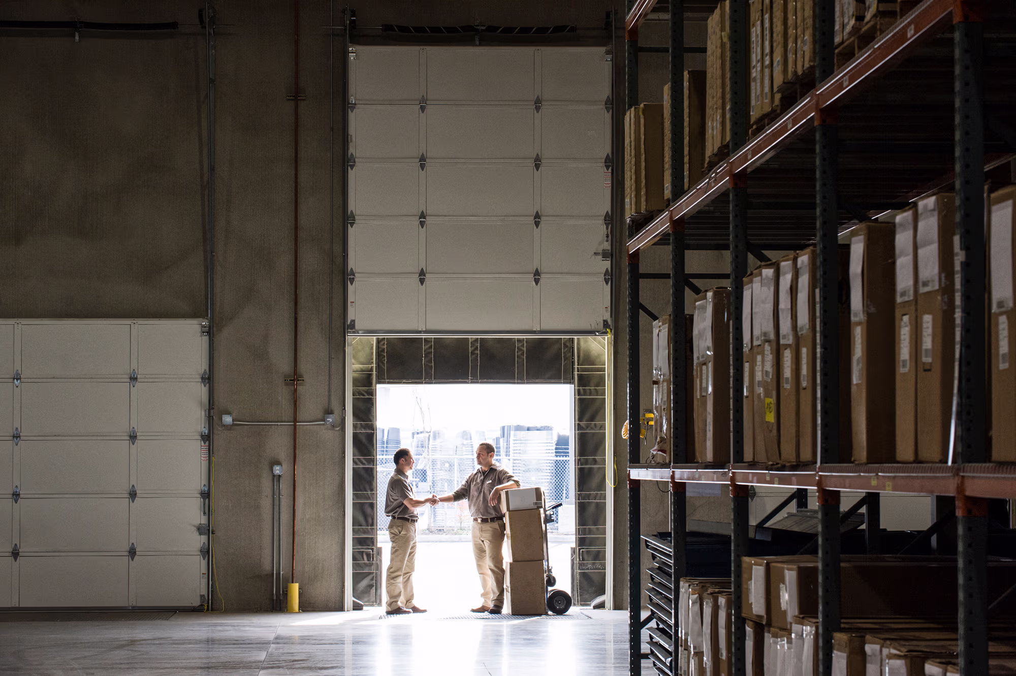 Two warehouse workers shaking hands near a loading dock doorway, with one standing next to a hand truck stacked with boxes and rows of shelving filled with cardboard boxes on the right side of the warehouse.