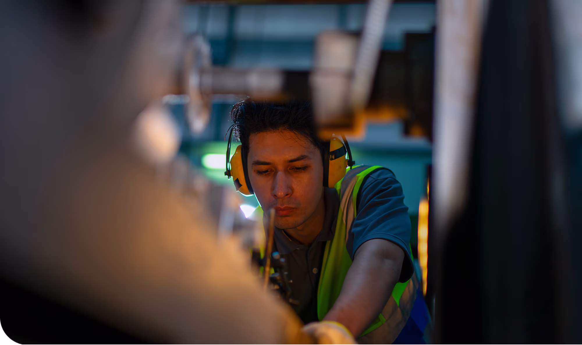 A male industrial worker wearing yellow ear protection and a green safety vest operates machinery in a dimly lit factory.