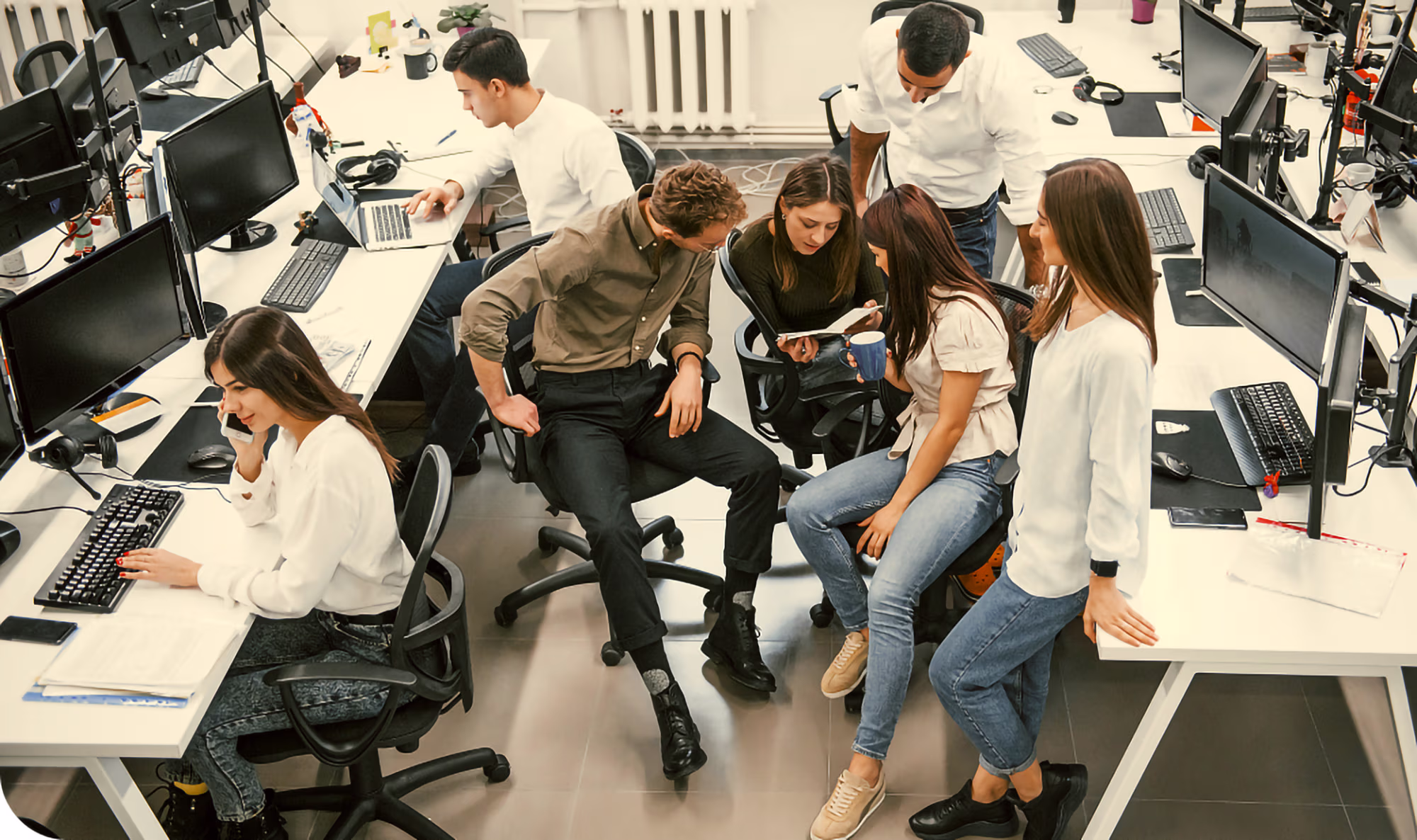 Six young business people in a modern office with multiple computer workstations. Two are seated working individually while four others gather closely, looking at a smartphone and a cup, engaged in conversation and collaboration.