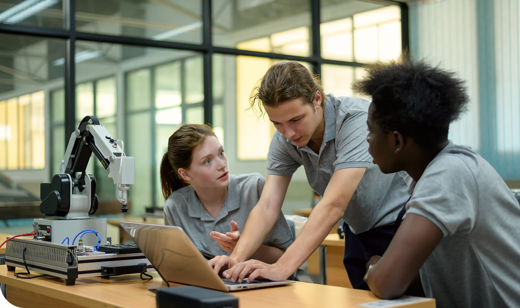 Three young adults in a modern classroom engaged in robotics learning, with one typing on a laptop, another explaining, and a robotic arm on the table.