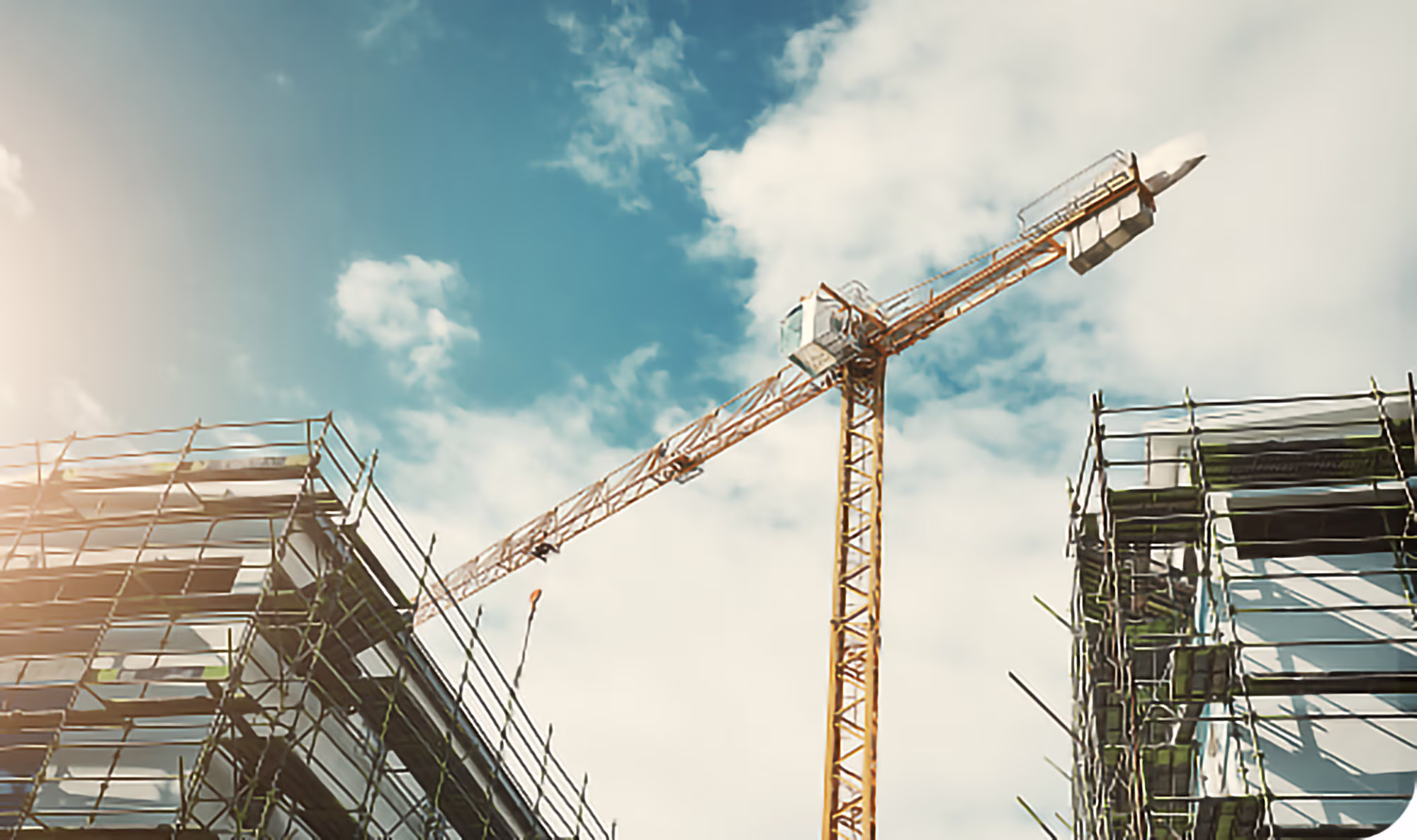 Yellow construction crane positioned between two buildings under construction with scaffolding, against a blue sky with clouds.