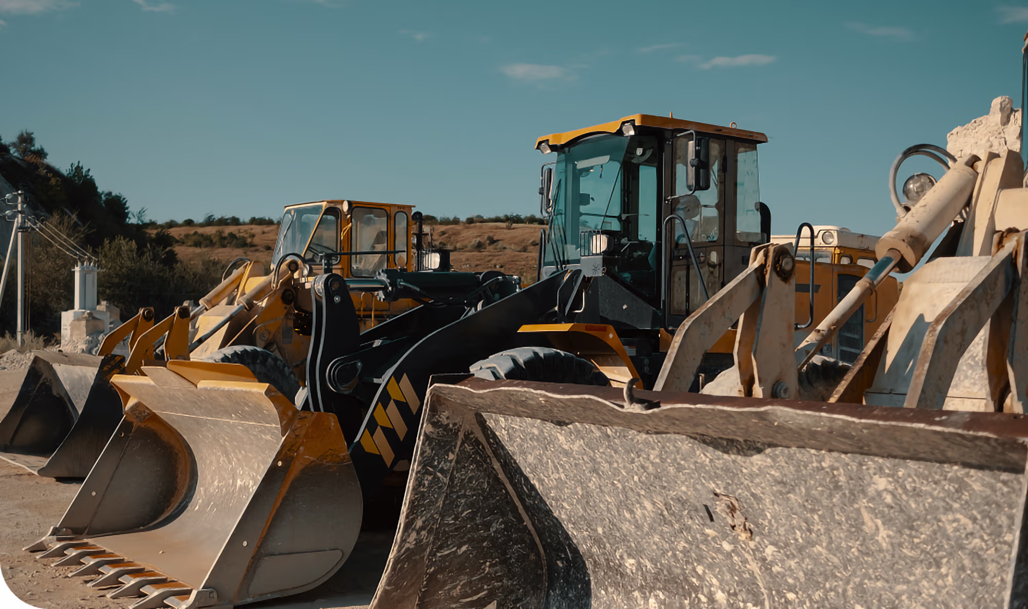 Several large yellow and black heavy construction machines with front loaders parked side by side on a dirt surface under a clear blue sky.