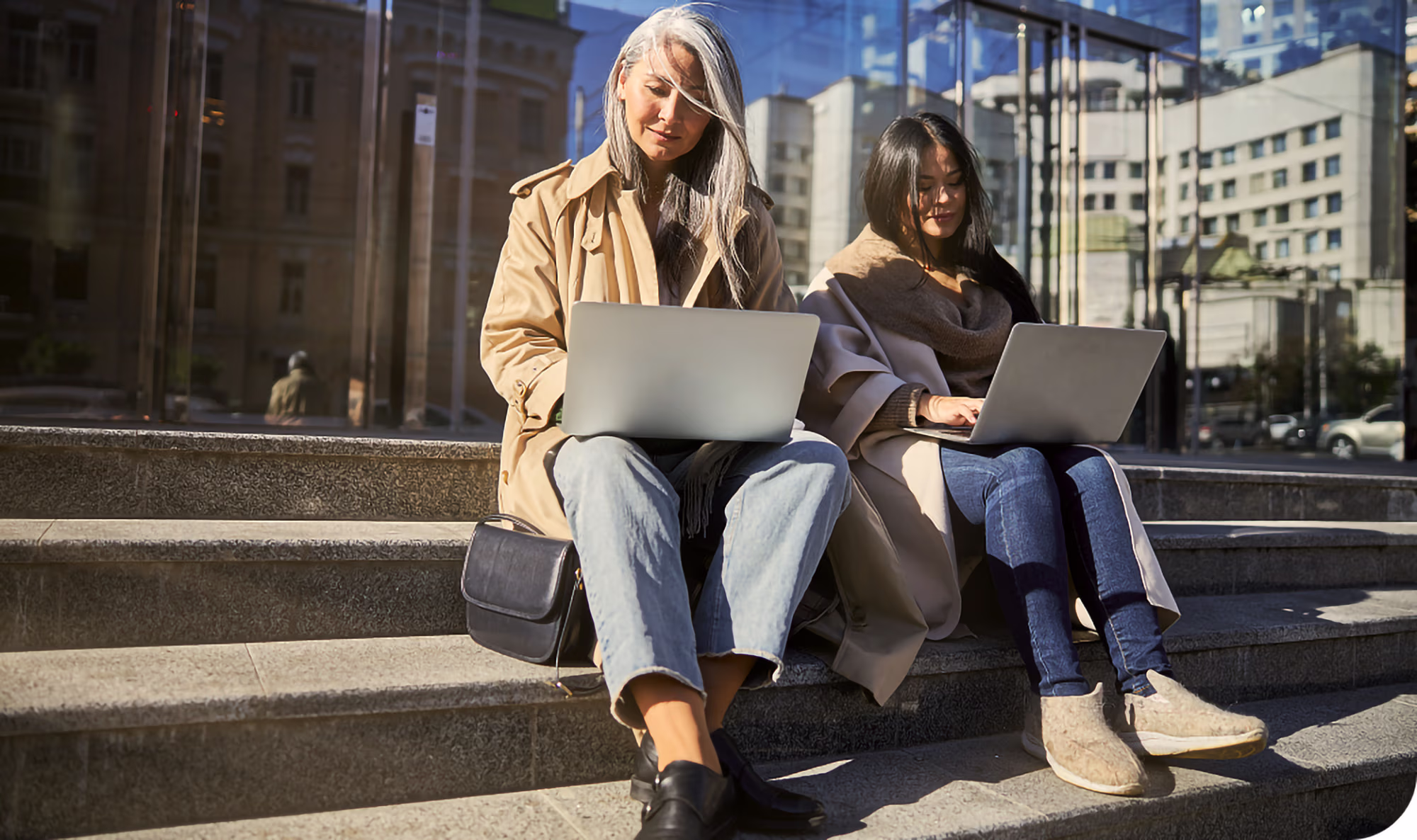 Two women sitting on outdoor concrete steps working on laptops, with reflections of buildings in the glass behind them.