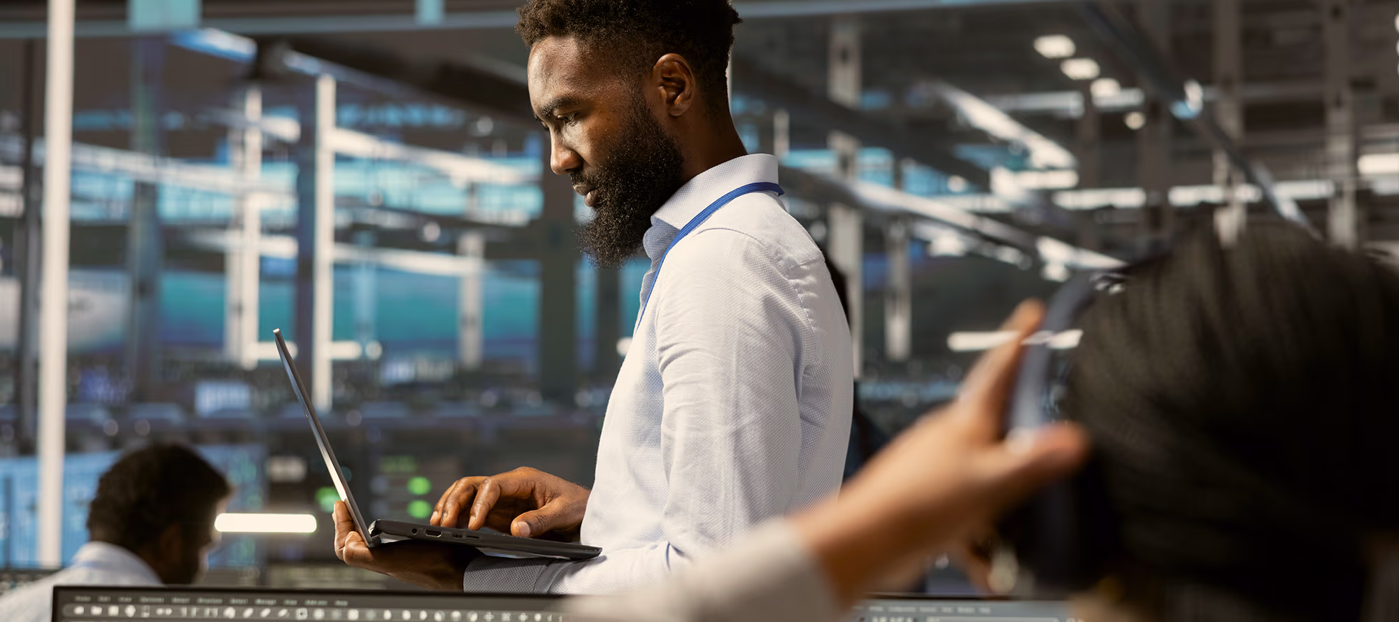 Man in a white shirt with a beard working on a laptop inside a data center with server racks and another person in the foreground.