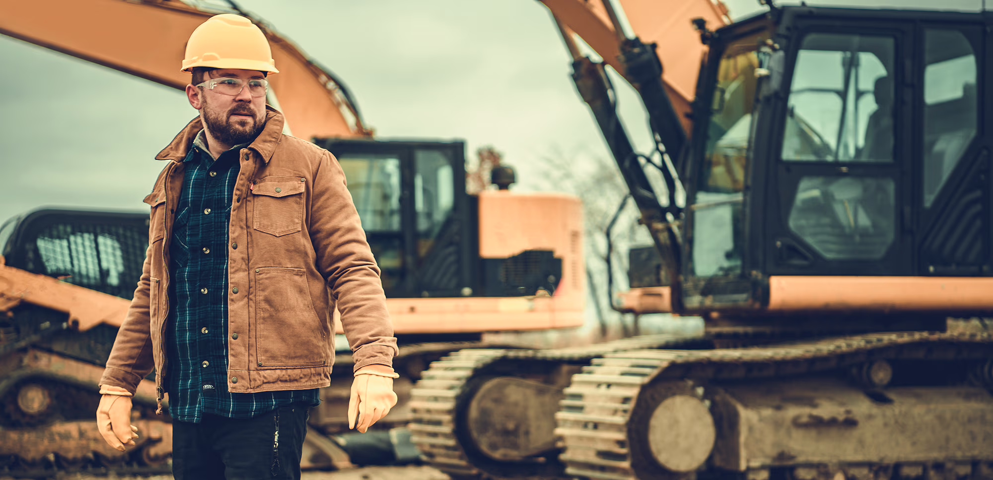 Construction worker wearing a hard hat, safety glasses, and brown jacket standing in front of heavy yellow excavators on a construction site.