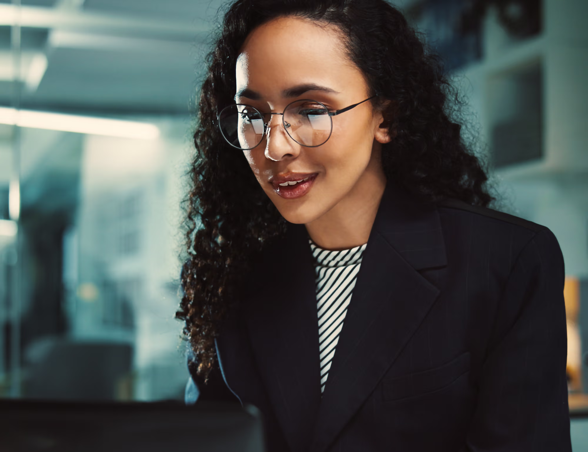 A woman with curly hair and round glasses looking at a computer screen in a modern office setting.