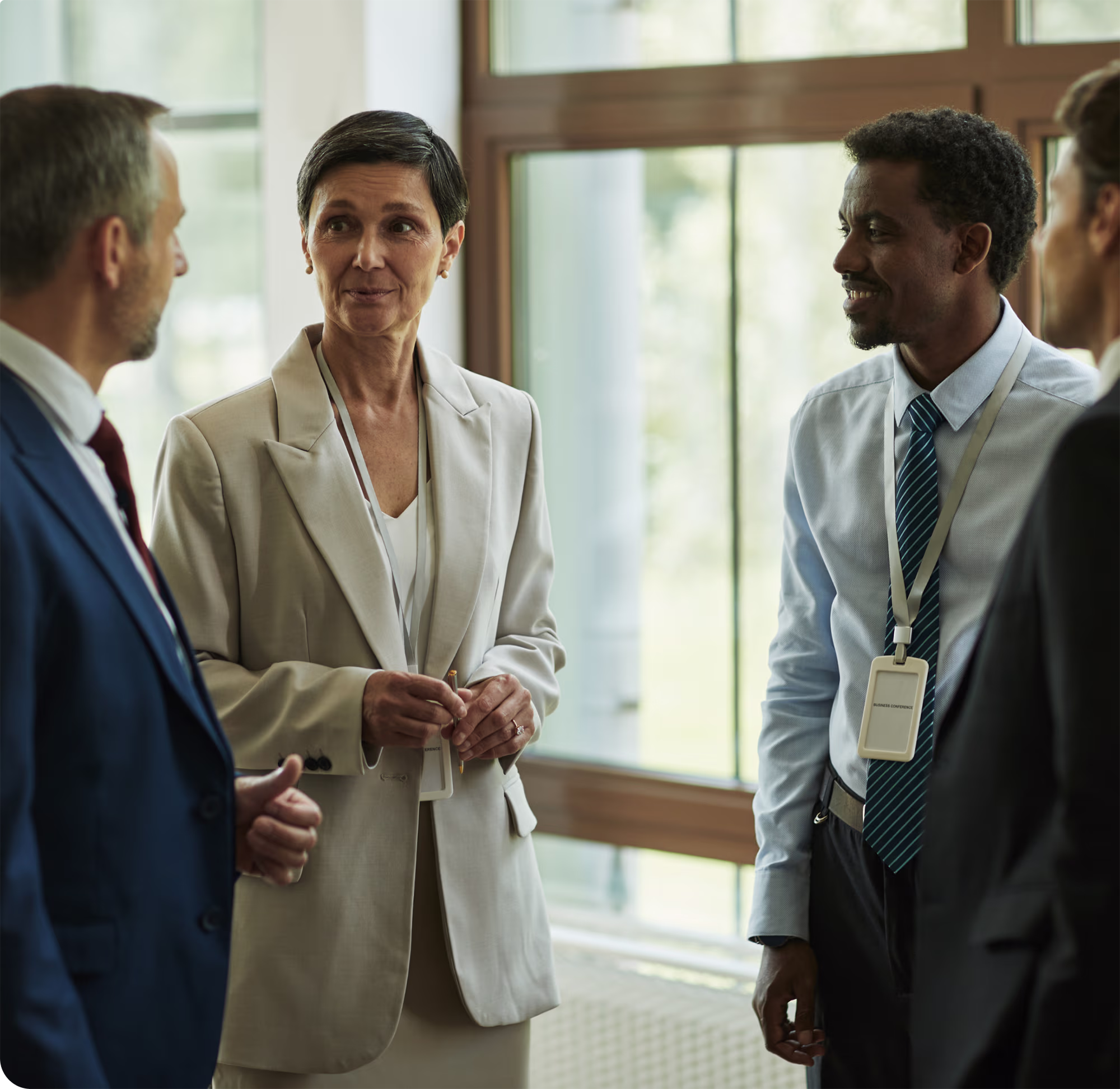 Four business professionals engaged in conversation in a bright office setting with large windows, two men wearing suits and two individuals wearing business attire with ID badges.
