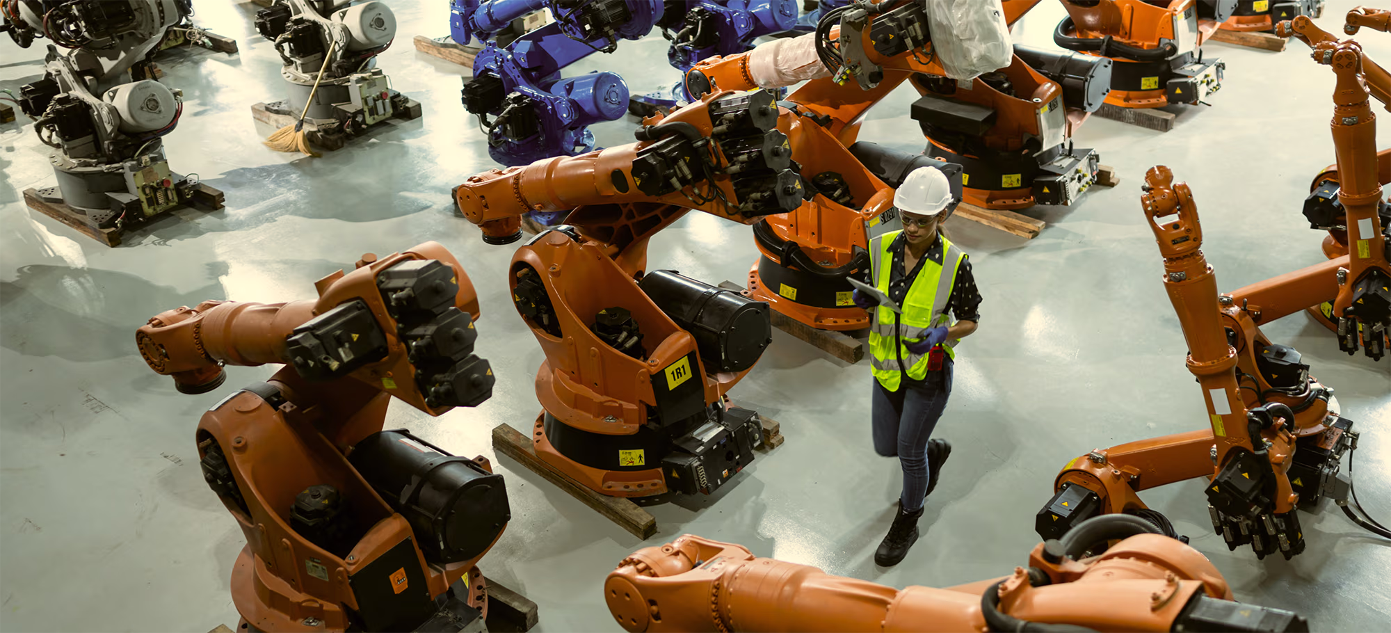 A worker in safety helmet and neon vest inspects multiple large orange industrial robotic arms arranged on a factory floor.