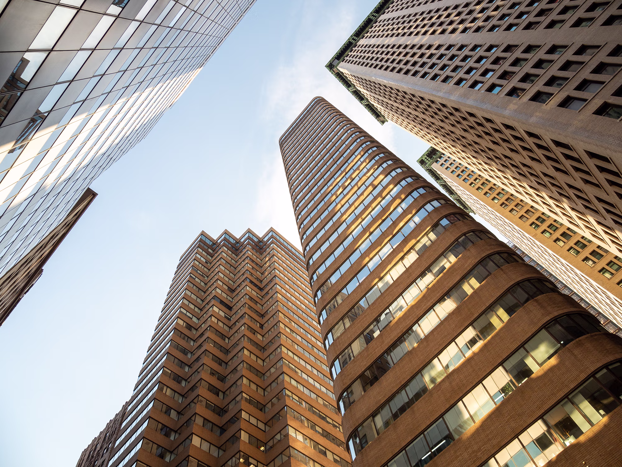 Low-angle view of multiple tall buildings with varied facades, including glass windows and brick patterns, against a light blue sky.