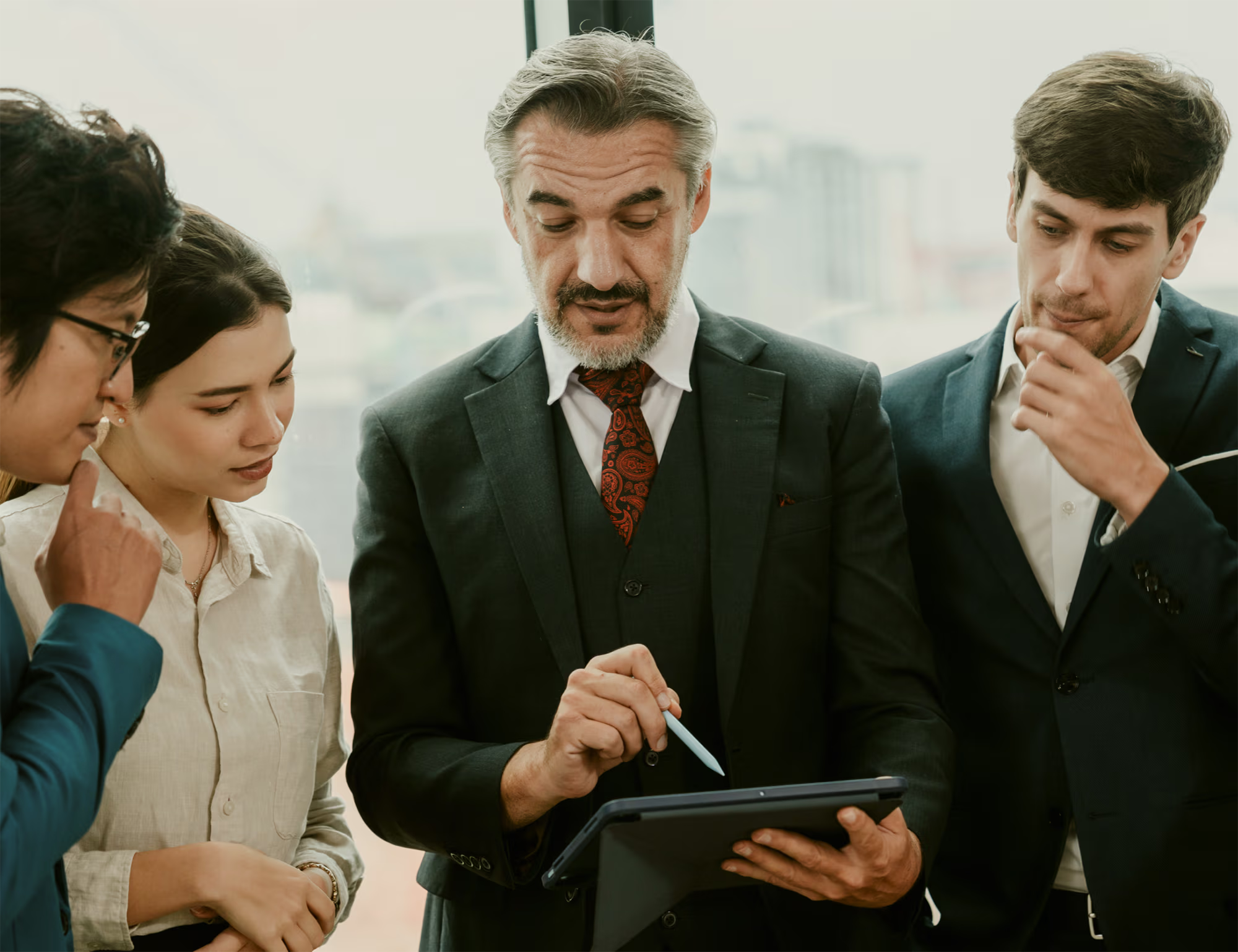 Four business professionals in formal attire gathered around a tablet, attentively looking at the screen as the man in the center uses a stylus to point or interact with it.