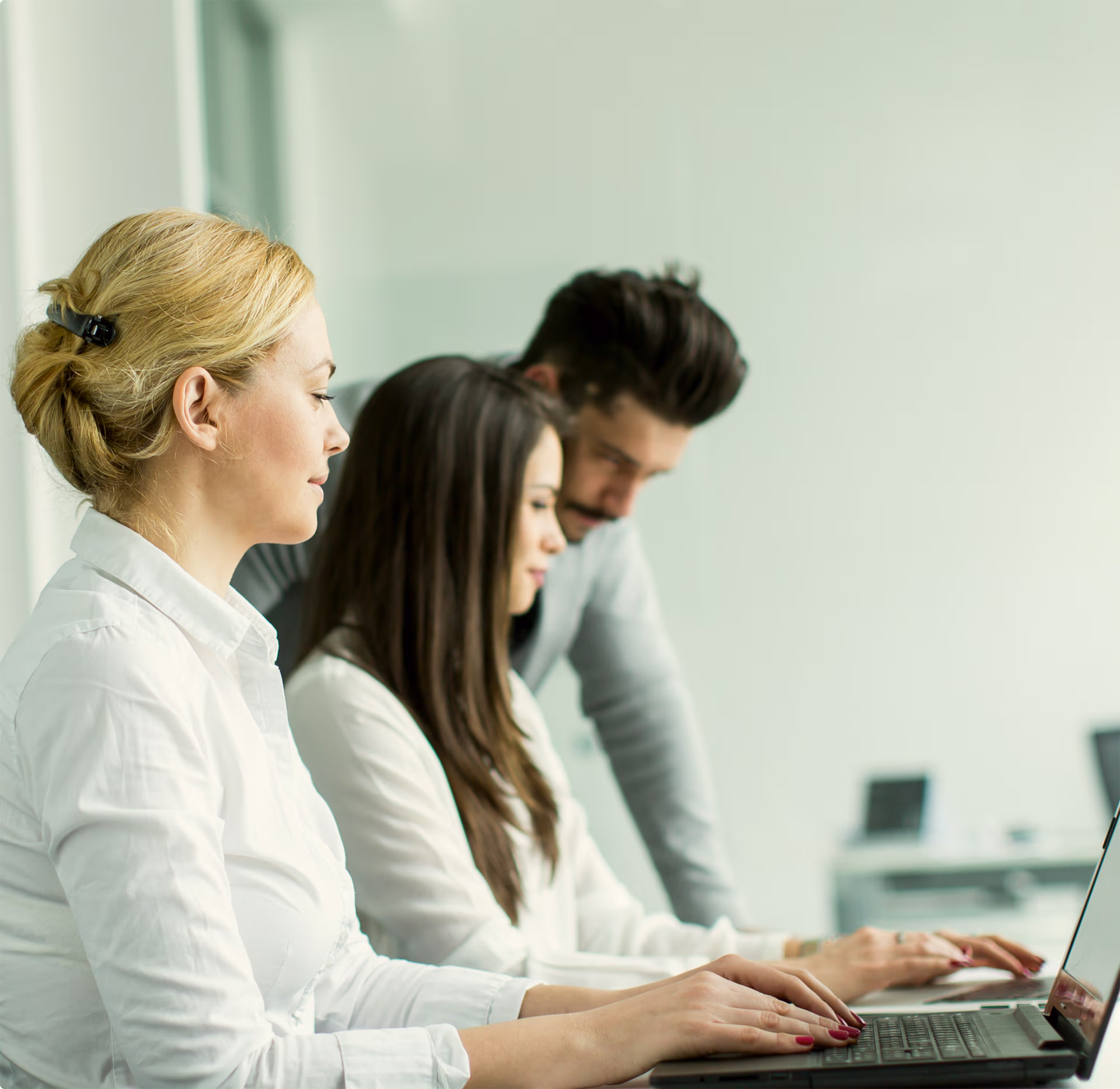 Three professionals working together using two laptops in a bright, modern office setting.