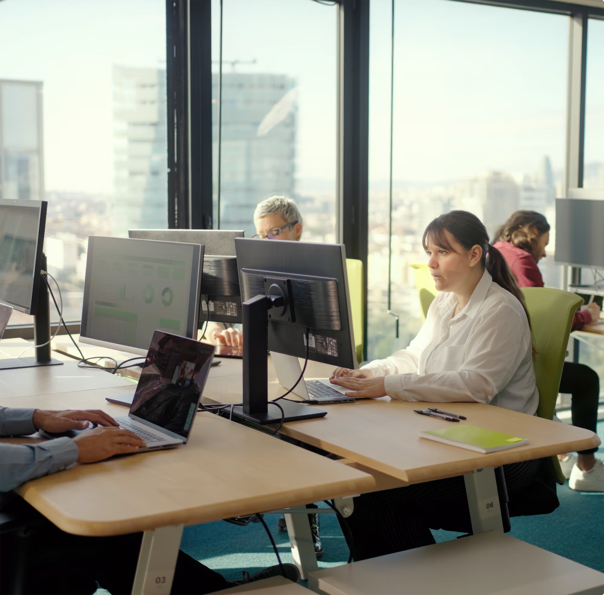 Office space with natural light where three people are working at desks with large windows showing a city view; one woman in white shirt types at a computer, a person using a laptop is partially visible, and a third person in red focuses on their work at a desk near a floor lamp.