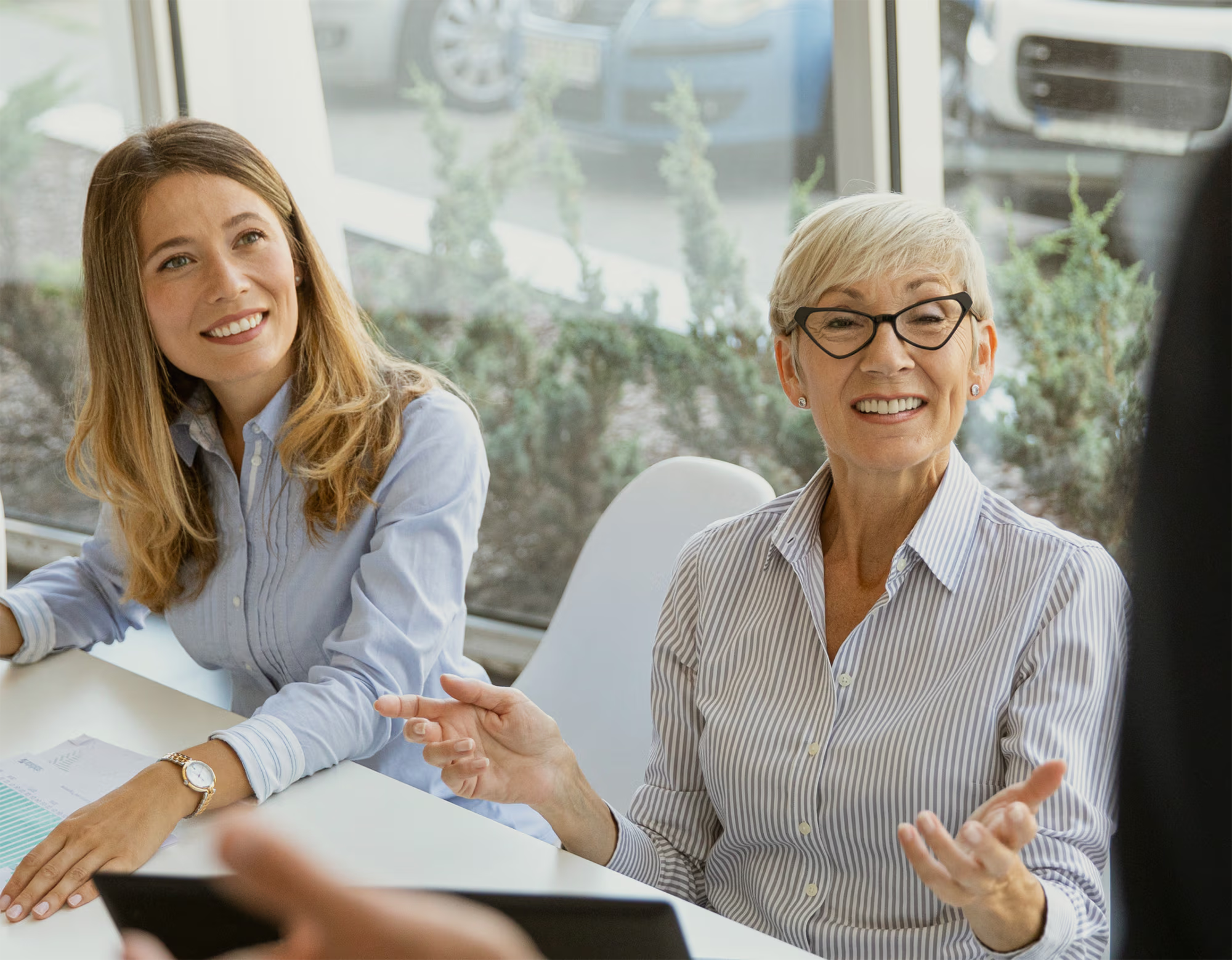 Two professional women sitting at a table in a bright office, smiling and engaged in conversation, one woman wearing glasses and a striped shirt.