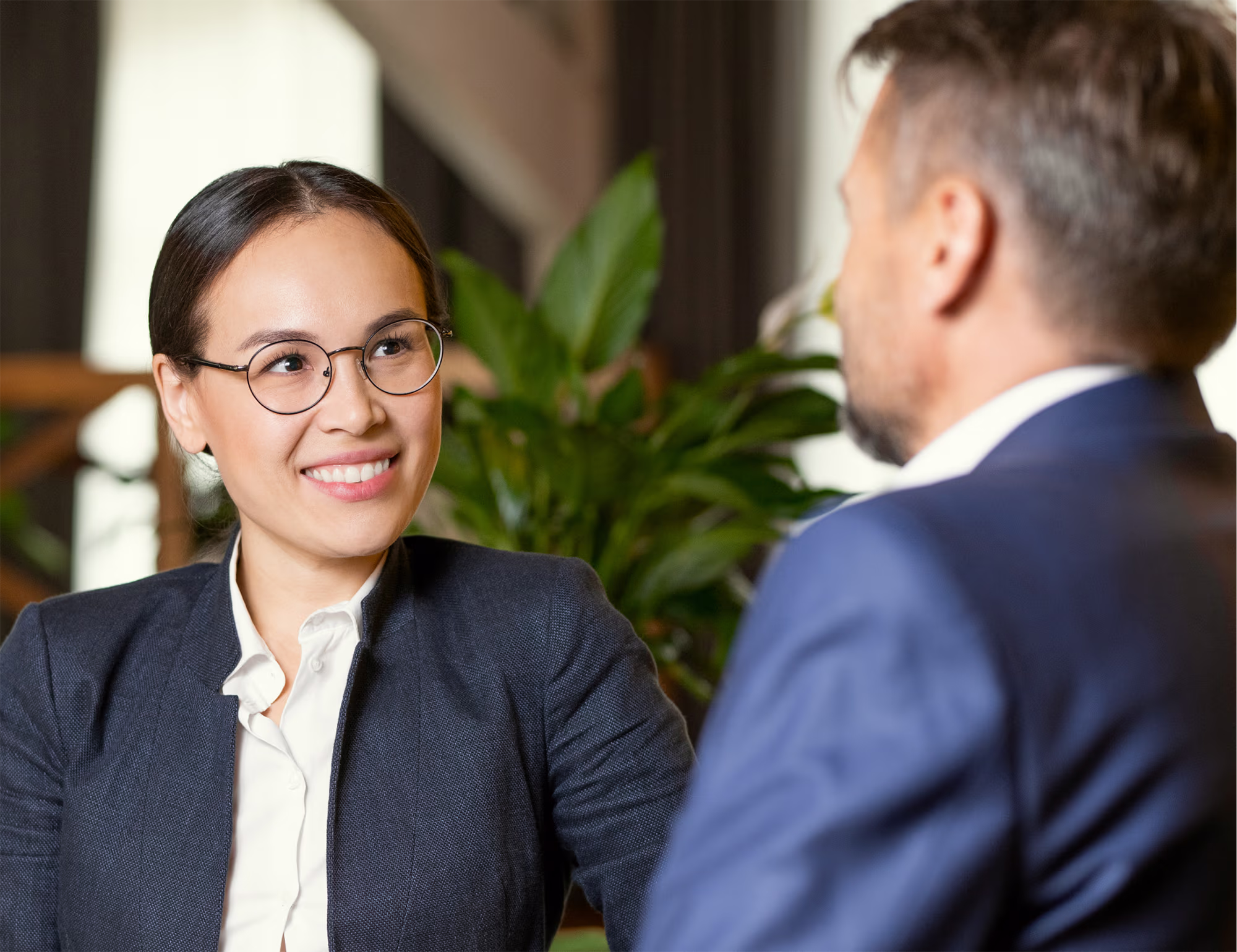 Two professionals having a conversation indoors with green plants in the background; a woman with glasses and a dark blazer smiles at a man in a blue suit.