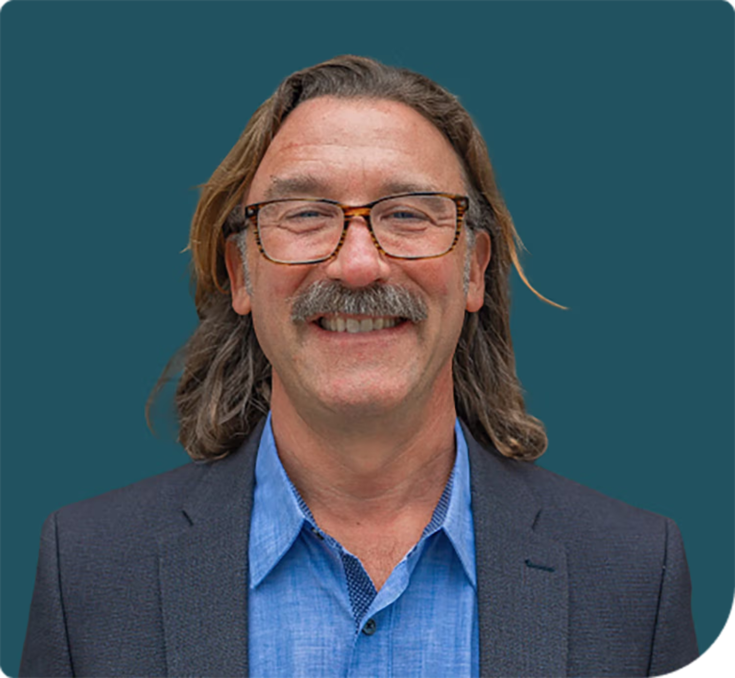 Smiling man with a mustache and shoulder-length hair wearing tortoiseshell glasses, a blue shirt, and a dark blazer against a teal background.