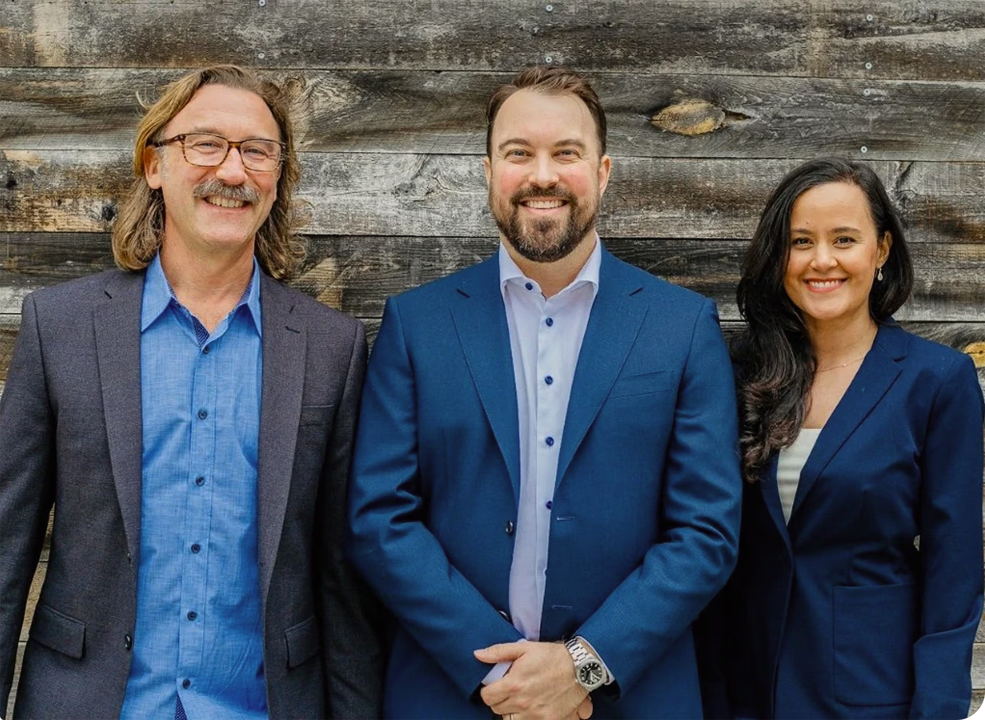 Three professionals smiling in front of a wooden wall, dressed in business attire: a man with glasses and long hair in a gray blazer and blue shirt, a man with a beard in a dark blue suit and white shirt, and a woman with long dark hair in a dark blue suit and white top.
