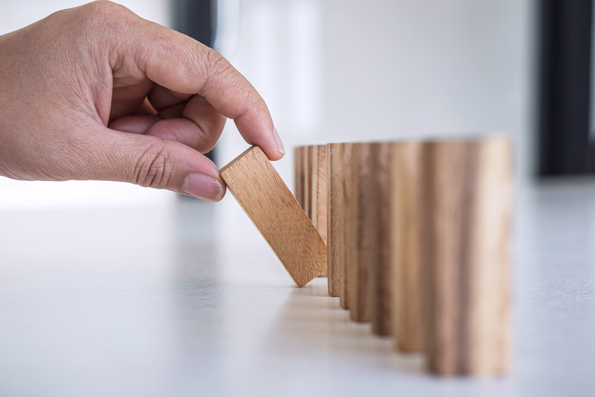 A hand placing a wooden domino piece upright in front of a row of standing wooden dominoes on a flat surface.