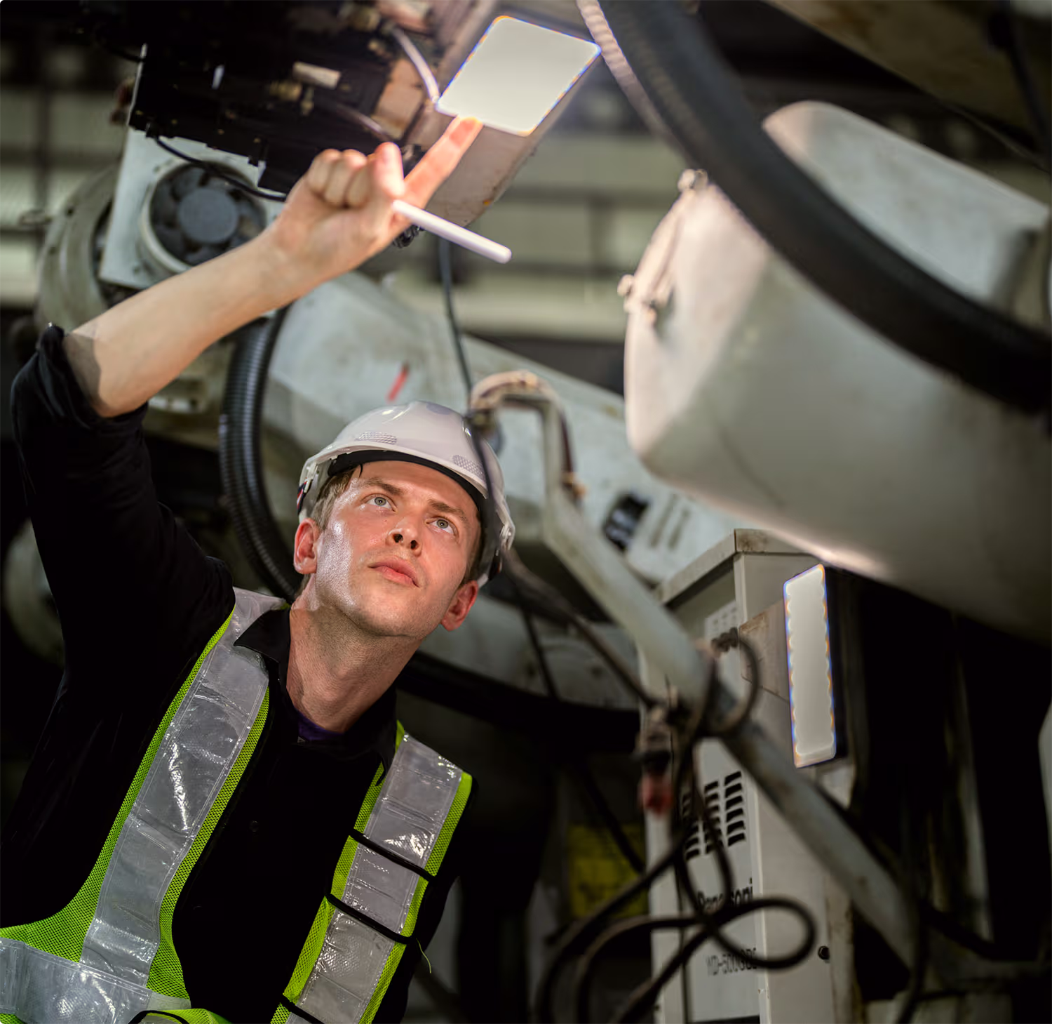Man wearing a white safety helmet and a reflective safety vest inspecting industrial machinery while pointing at a lit panel above him.