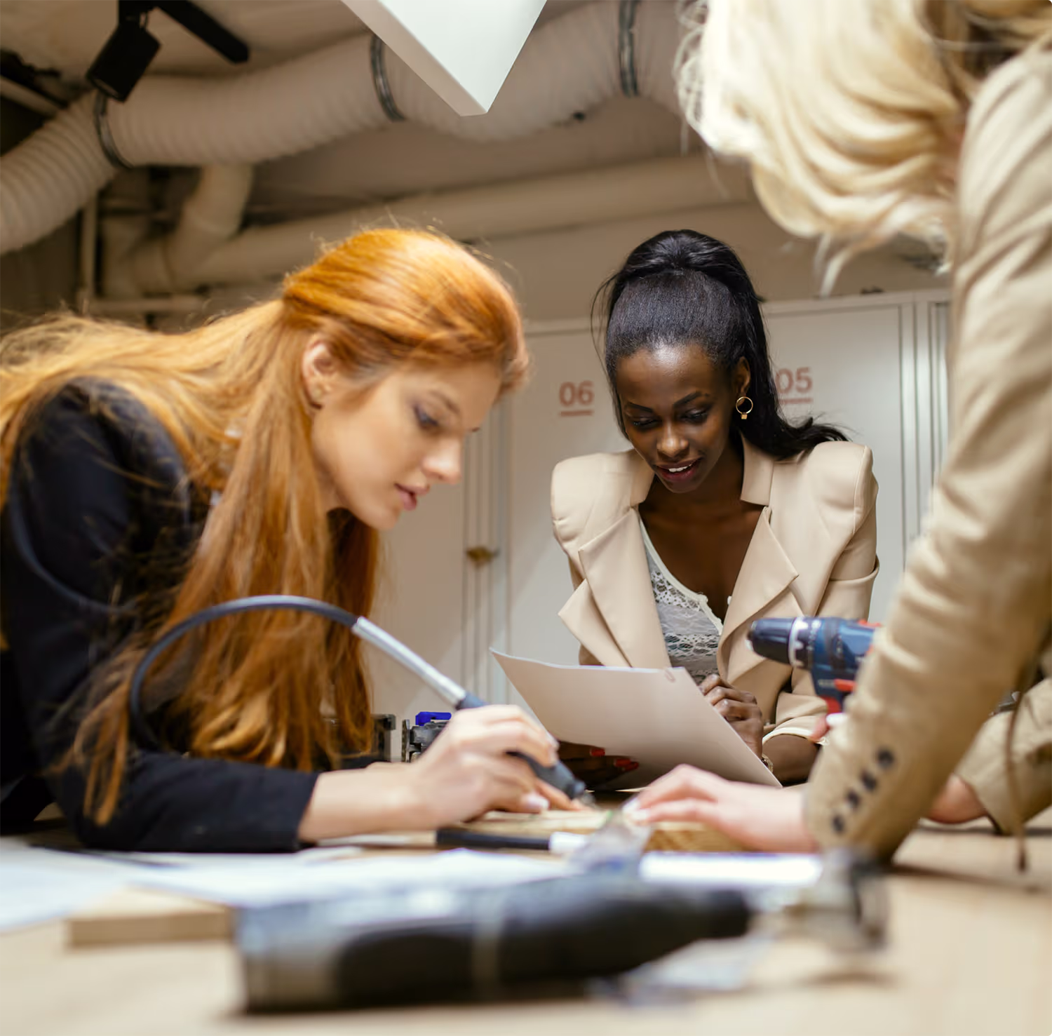 Three women collaborating over a workbench with tools; one with red hair is focused on a project using a tool, a dark-skinned woman with a ponytail and beige blazer is reading a document, and a blonde woman leans over the table.