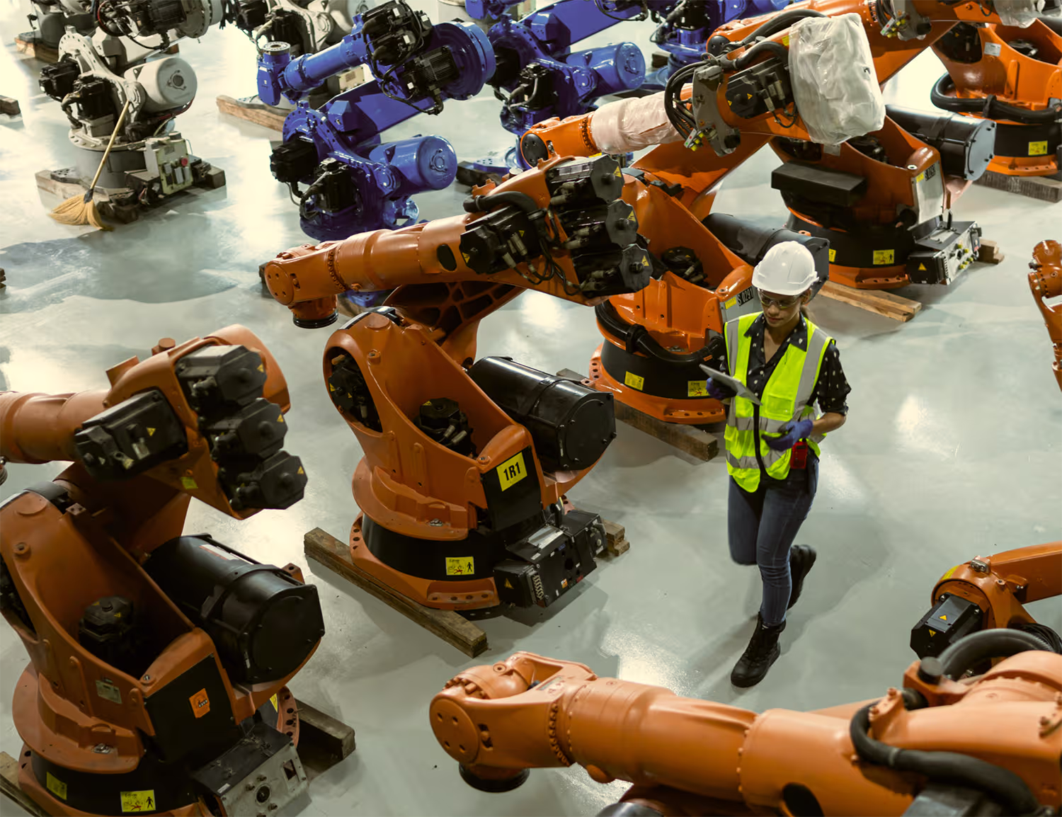 Woman wearing a white hardhat and yellow safety vest walking through a factory floor filled with large orange and blue industrial robotic arms.