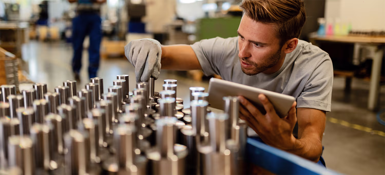 A man wearing a gray shirt and a glove inspects and arranges metal cylindrical parts on a table in a workshop while holding a tablet.