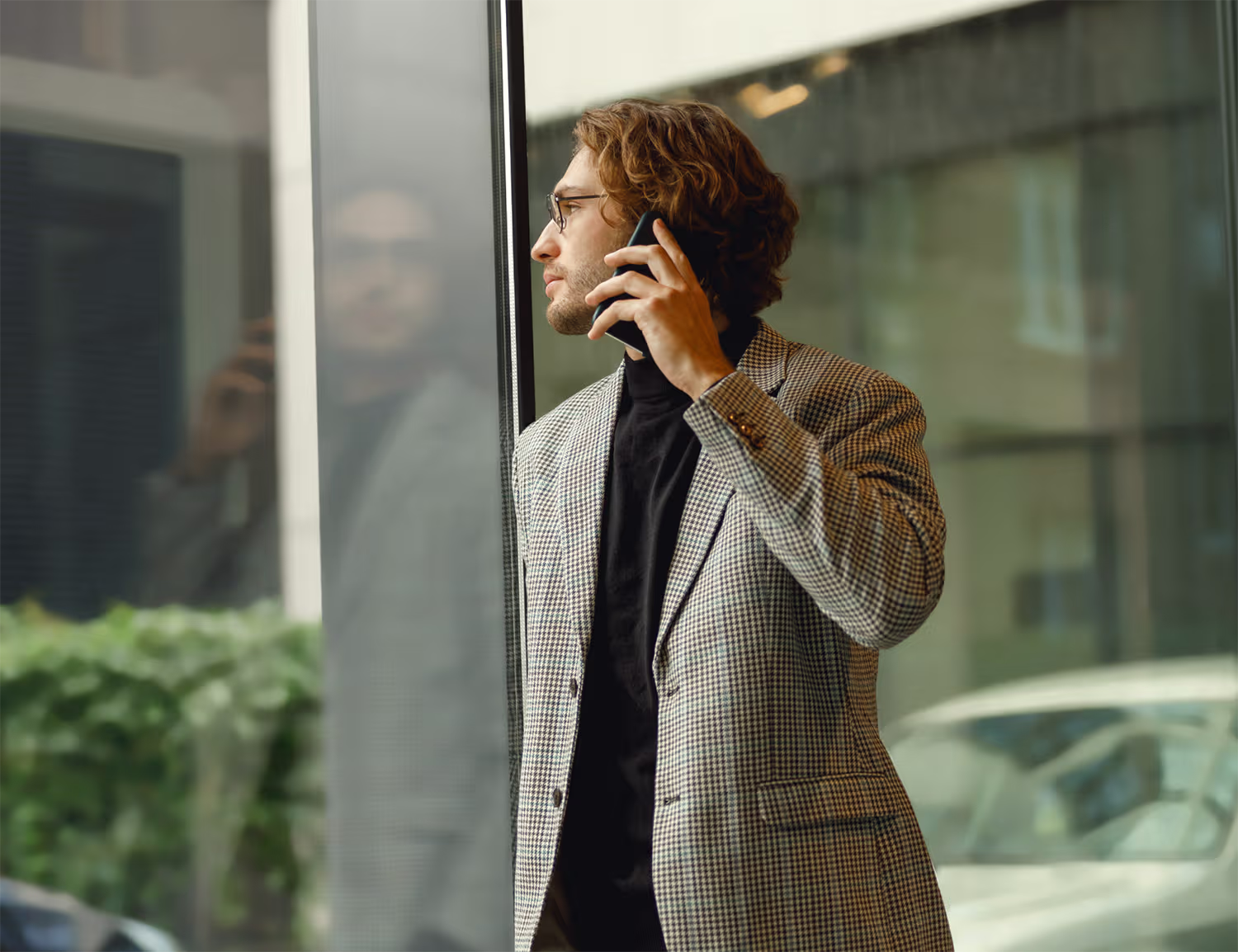 Man with curly hair and glasses wearing a checkered blazer and black shirt, standing indoors near a large window, holding a smartphone to his ear and looking outside.