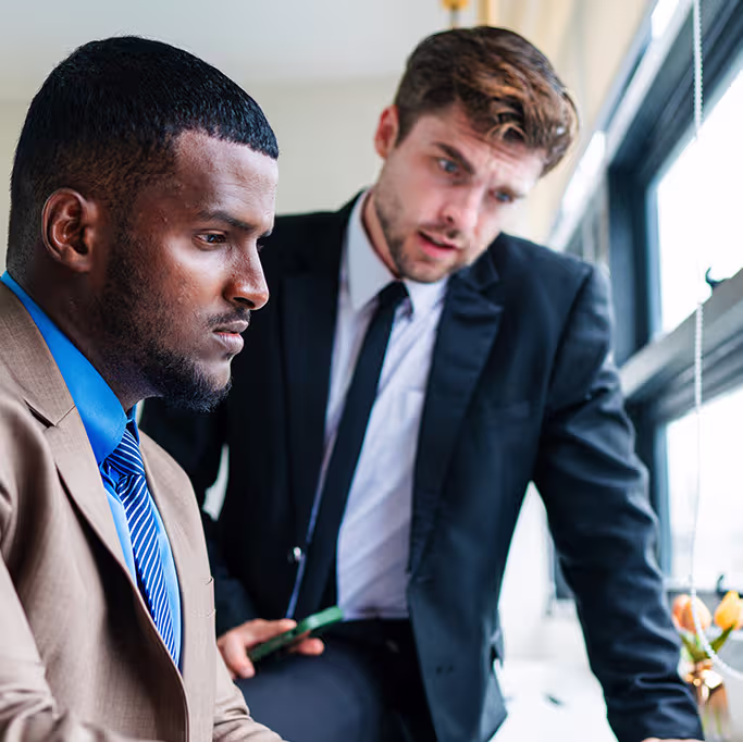 Two businessmen in suits engaged in a discussion by a window, one seated and focused, the other standing and looking intently.
