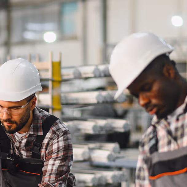 Two factory workers wearing white hard hats and plaid shirts with overalls, focusing on their tasks in an industrial setting with stacked metal rods in the background.