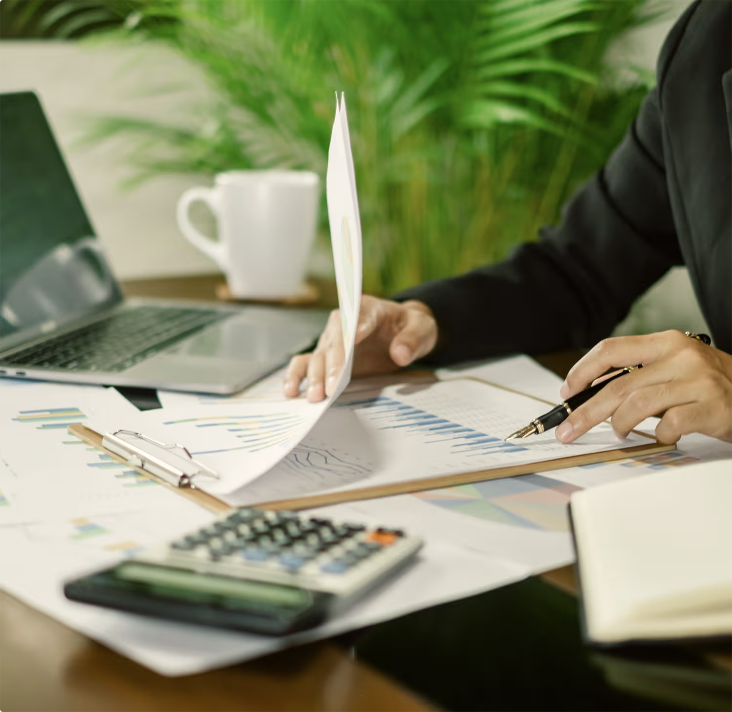 Person reviewing financial documents with charts and graphs on a desk, holding a pen and flipping a paper, with a calculator, laptop, coffee mug, and notebook nearby.