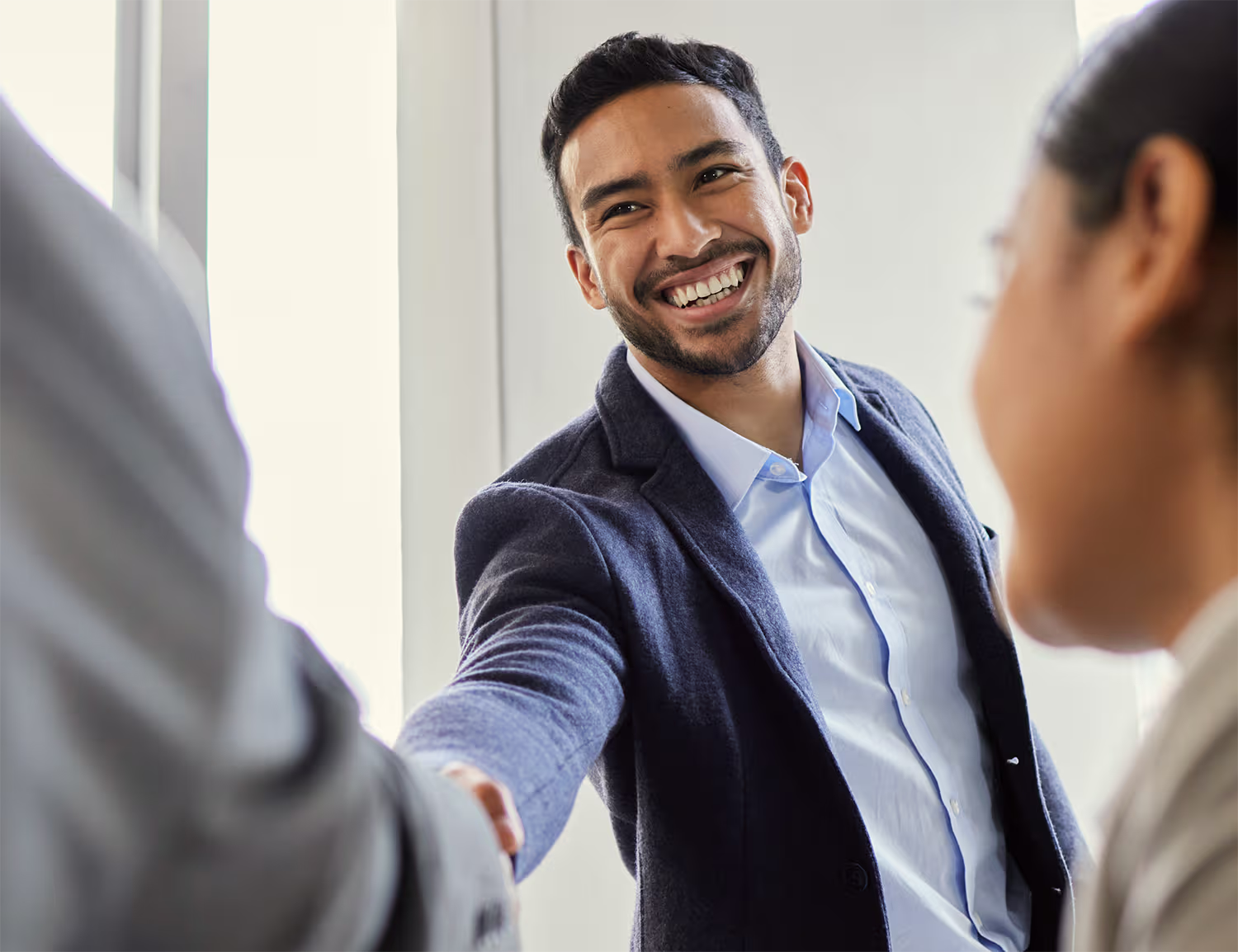 Smiling man in business attire shaking hands with another person during a meeting, with a woman facing him in the foreground.