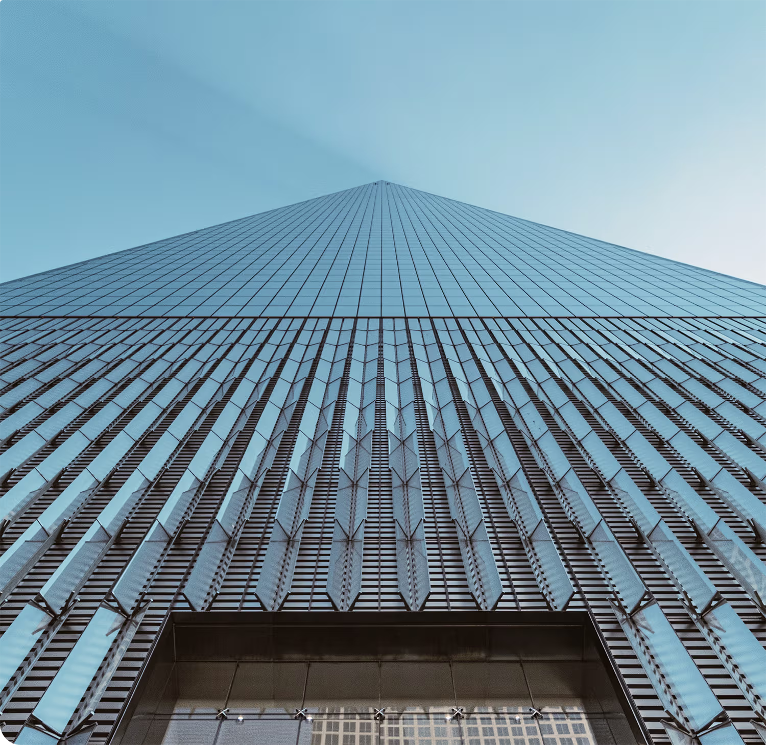Wall of a modern skyscraper with reflective glass panels converging towards a vanishing point in a clear blue sky, viewed from ground level looking up.