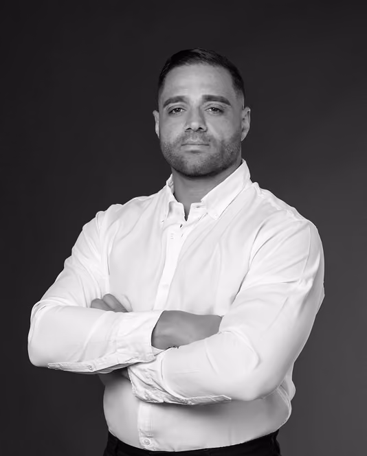 Man with short hair and trimmed beard wearing a white buttoned shirt with arms crossed.