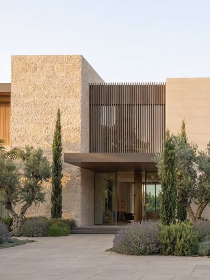 Modern house exterior with beige stone walls, vertical wooden slats on the upper level, a flat overhang above the glass entry door, and landscaped garden featuring tall cypress trees, olive trees, and lavender bushes along a stone-tiled driveway.