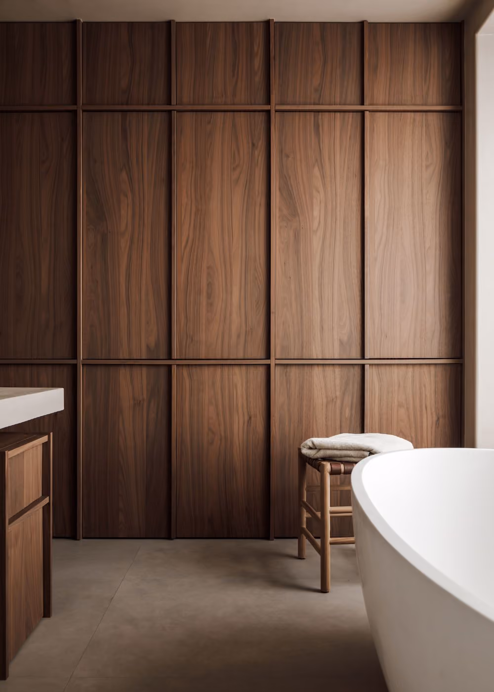 Modern bathroom with smooth wooden paneled wall, a white freestanding bathtub on the right, and a small wooden stool with folded towels.